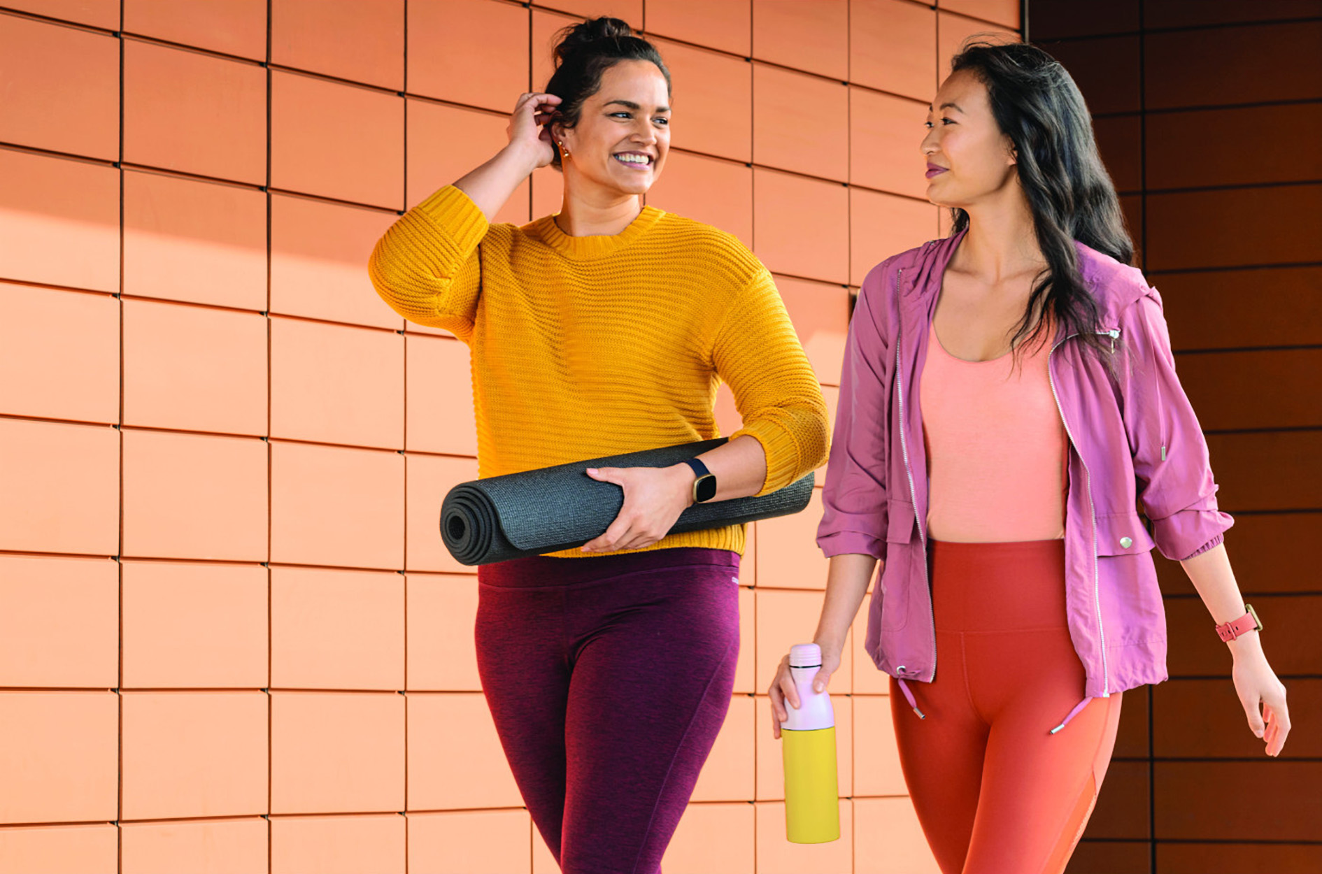Zwei Frauen gehen im Freien. Eine hält eine Yogamatte, die andere eine Wasserflasche. Beide lächeln.
