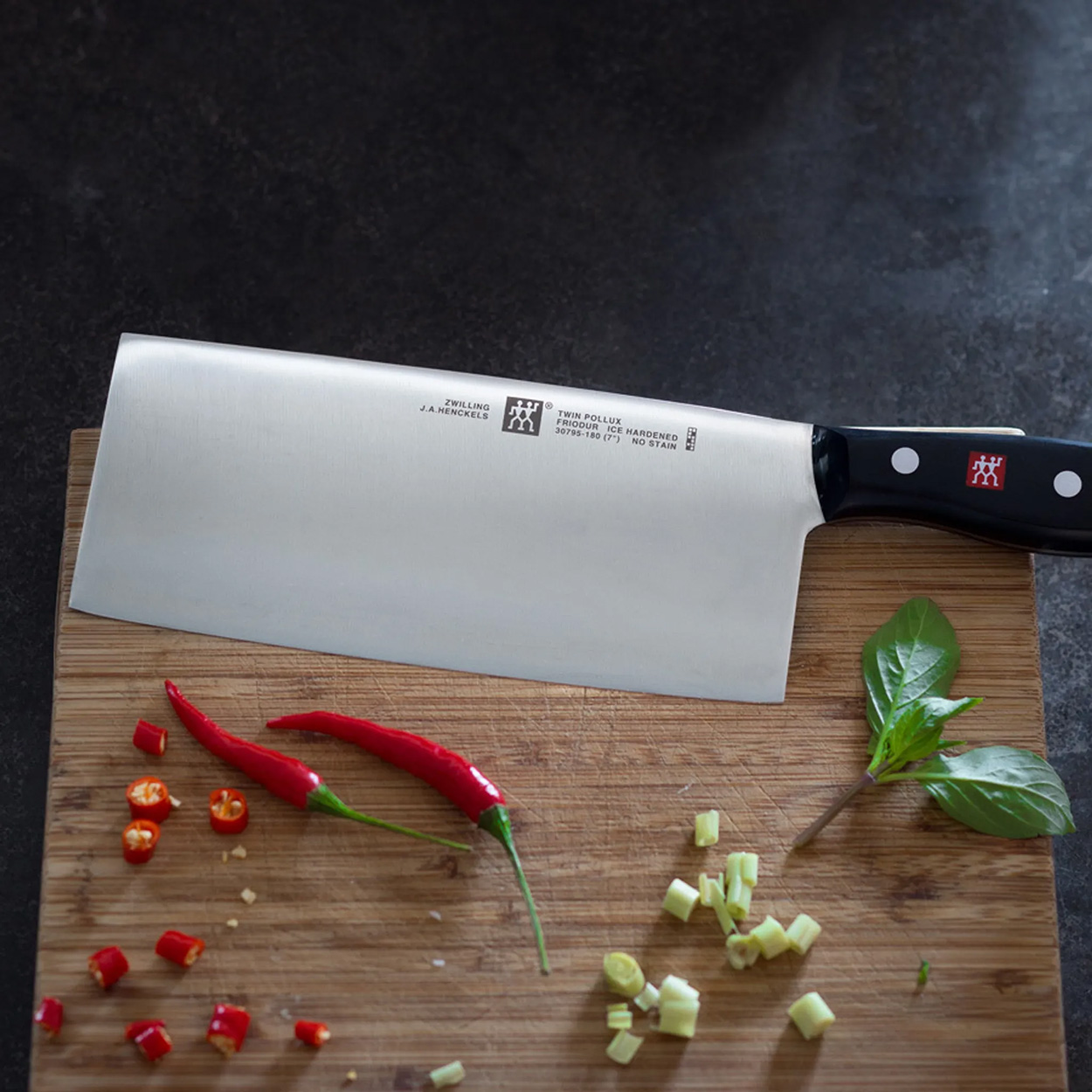 Großes Küchenmesser auf Holzbrett, mit roten Chilis und Kräutern. Dunkler Hintergrund.