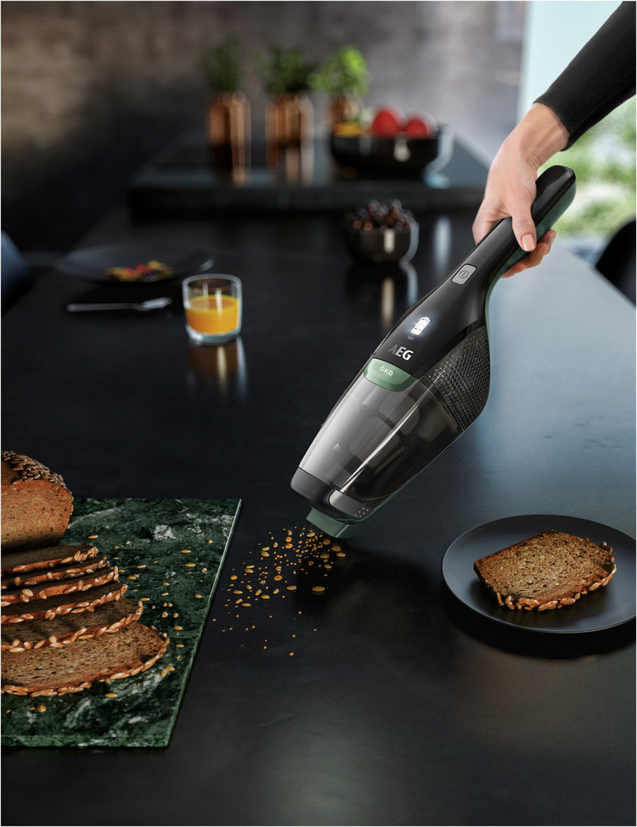 Eine Hand, die Krümel von einem schwarzen Tisch absaugt, mit Brot und Orangensaft im Hintergrund.