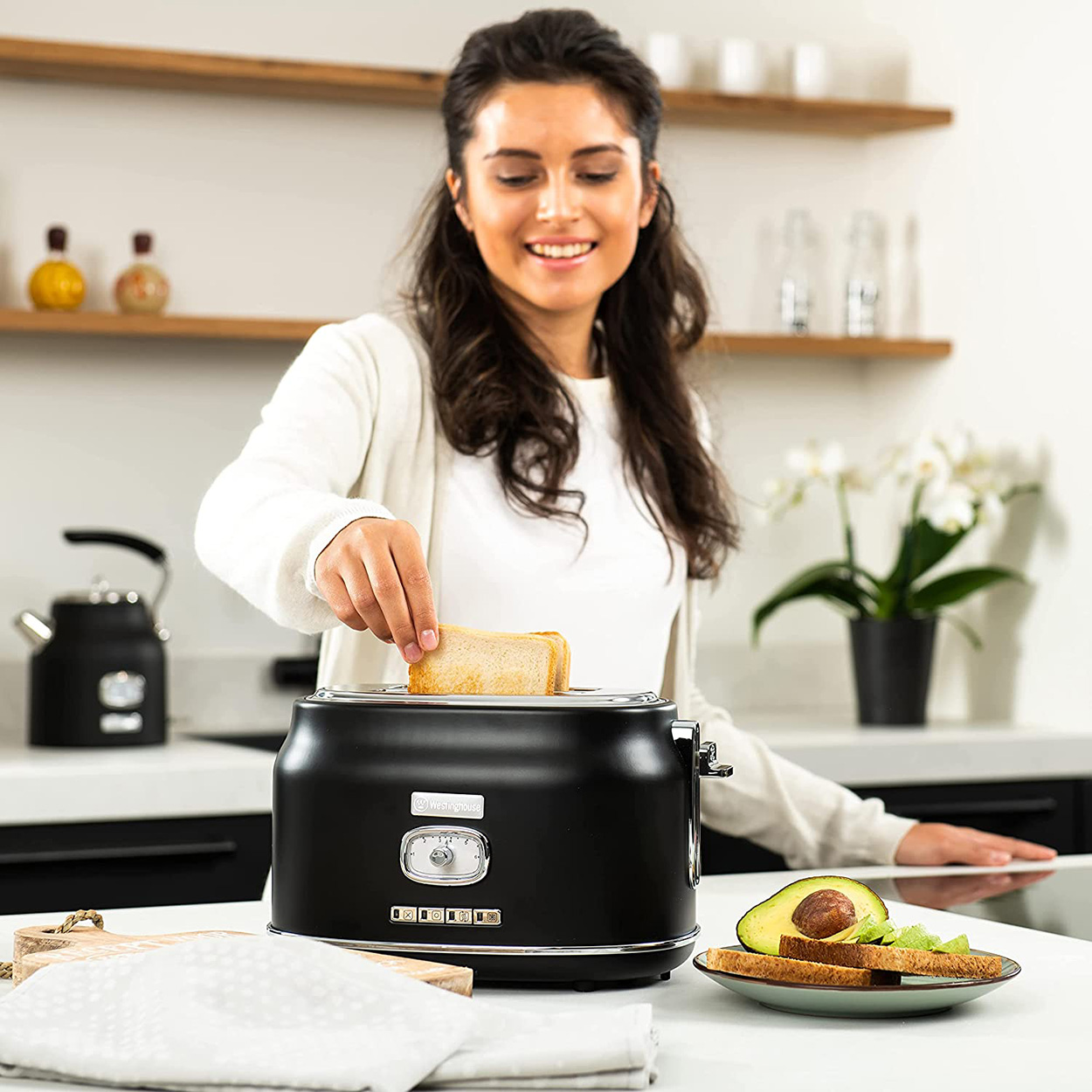 Frau legt Brot in einen schwarzen Toaster. Küche mit Avocado und Wasserkocher im Hintergrund.