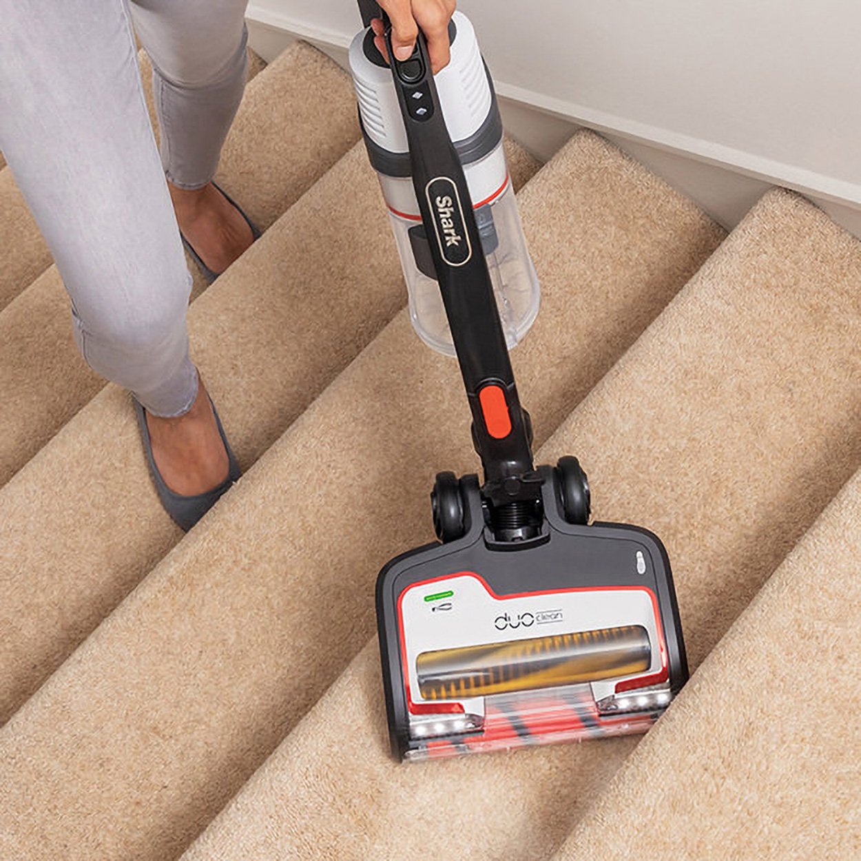 Una persona aspira escaleras alfombradas beige con una aspiradora Shark. La aspiradora es negra y roja.