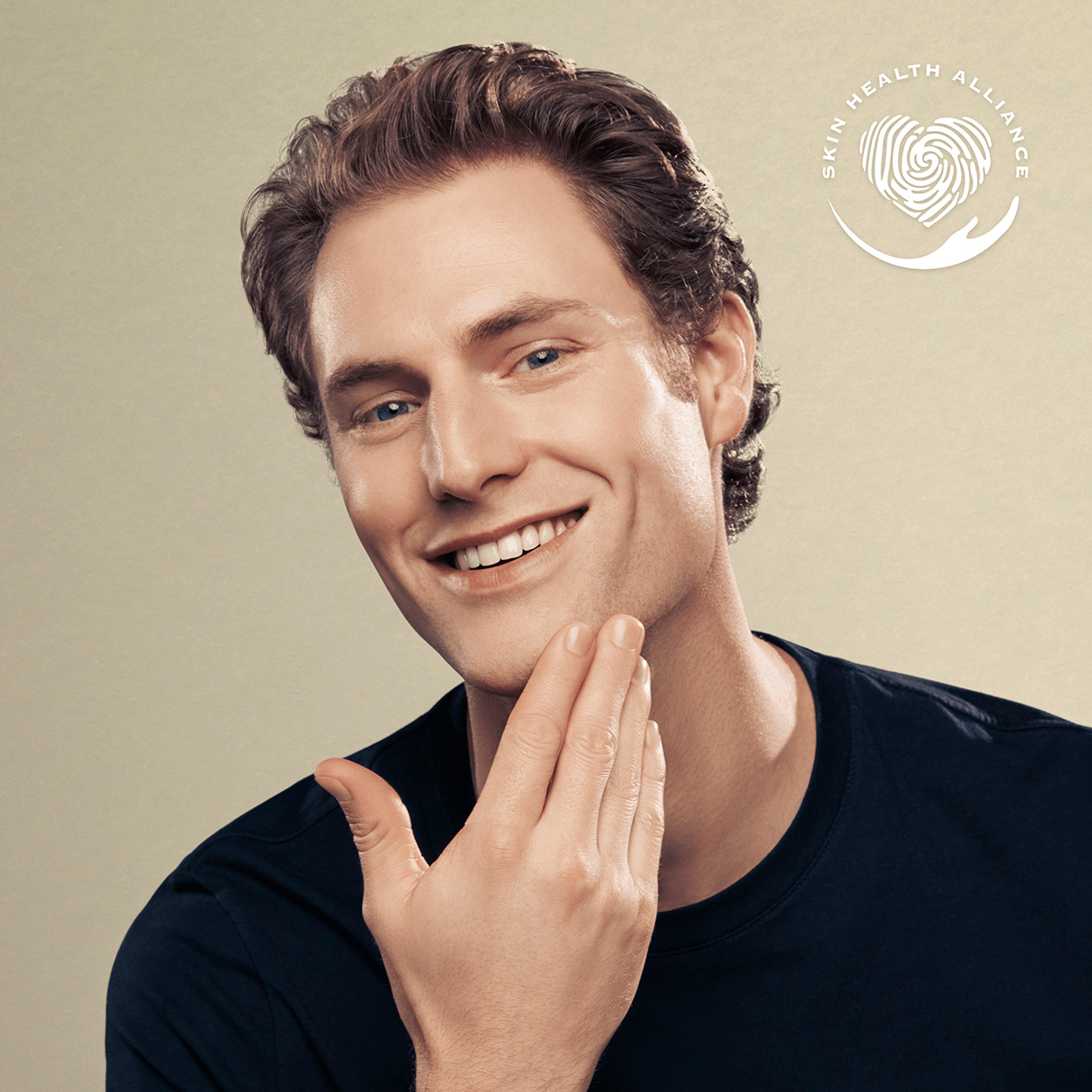 Un uomo con i capelli castani sorride e si tocca la guancia. Visibile un logo Skin Health Alliance.