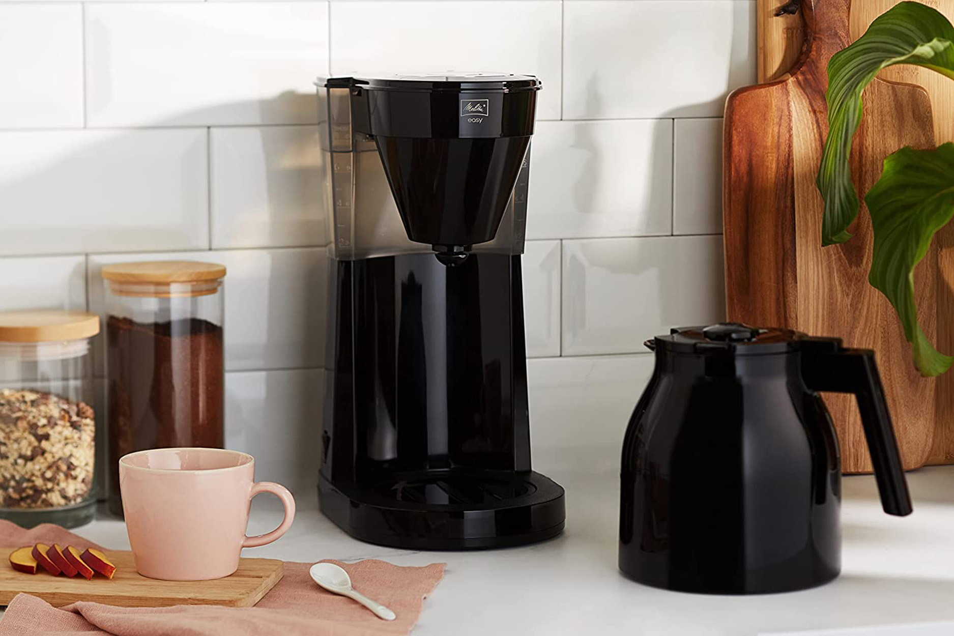 Czarny ekspres do kawy z karafką, różowy kubek i słoik z ziarnami kawy. Drewniane deski do krojenia w tle.