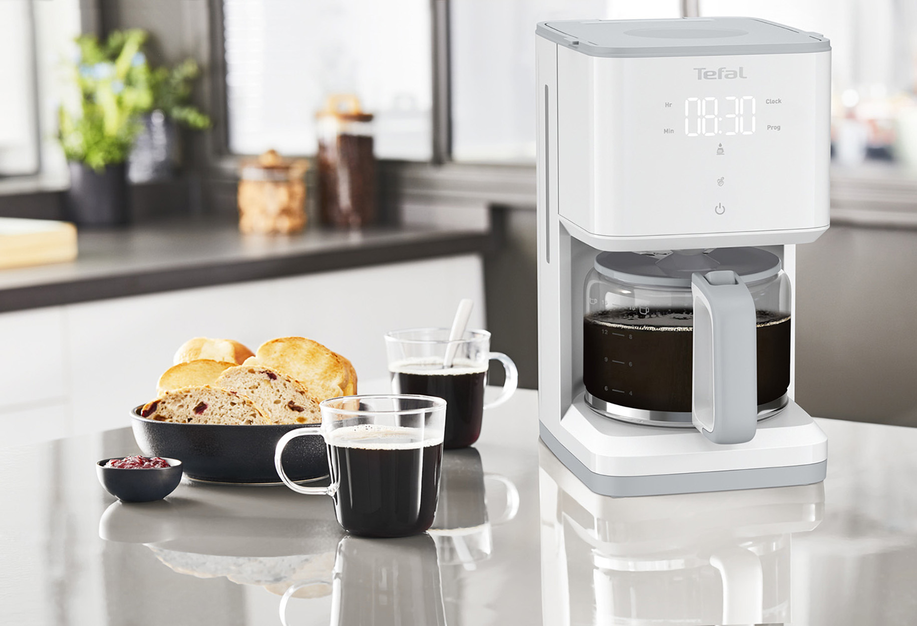 Biały ekspres do kawy ze szklaną karafką, kubkami i jedzeniem na blacie kuchennym.