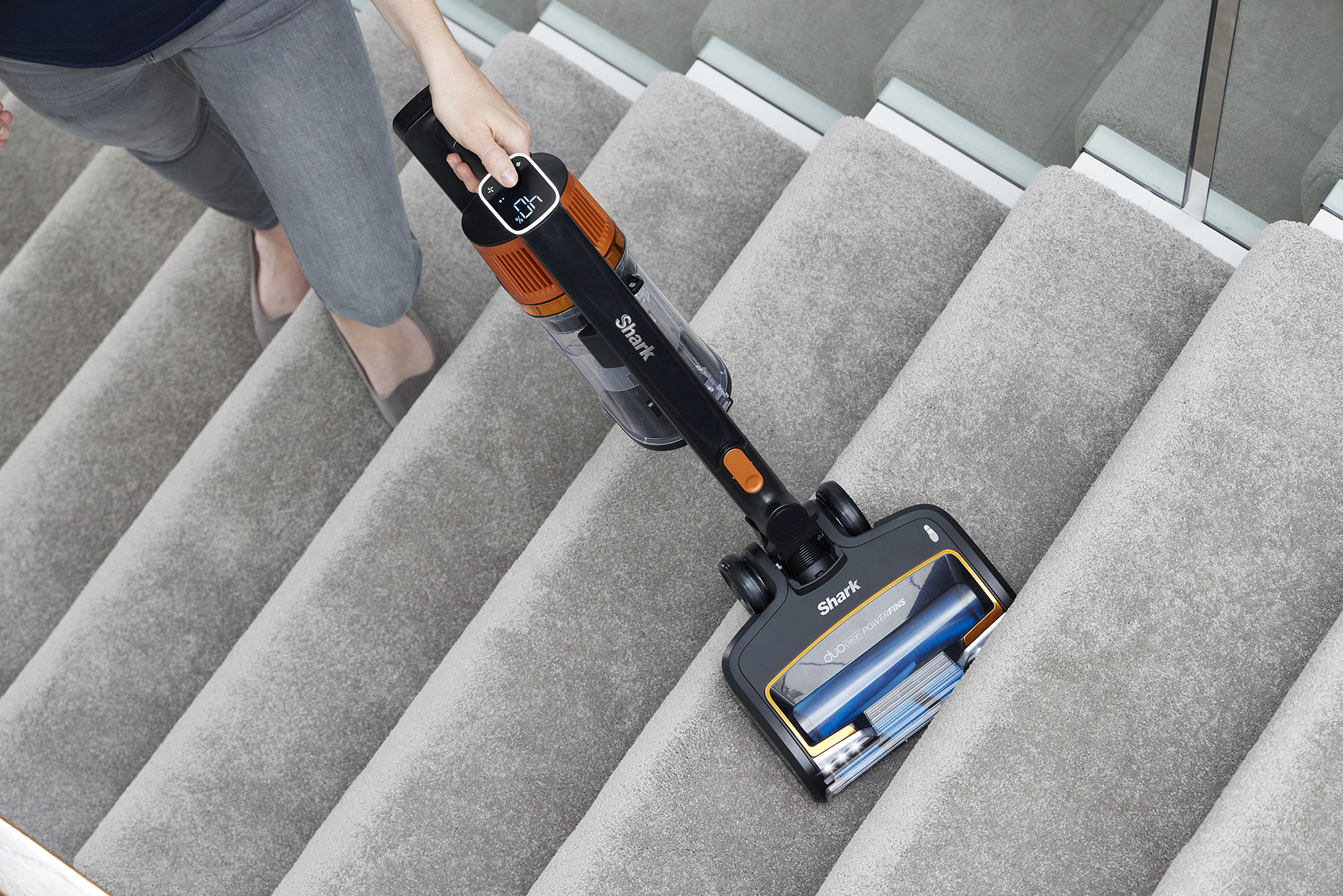 Persona aspirando escaleras alfombradas con una aspiradora Shark negra y naranja. Escaleras y alfombra grises.