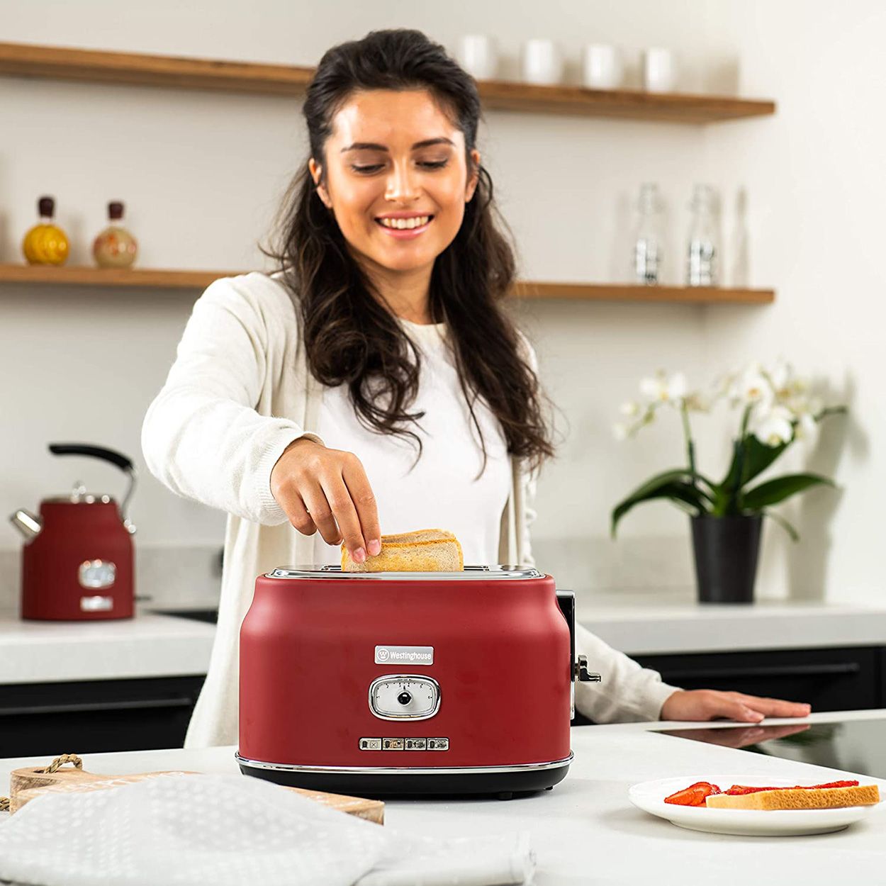 Frau legt Brot in einen roten Toaster auf eine weiße Theke, mit einem Wasserkocher und Blumen.