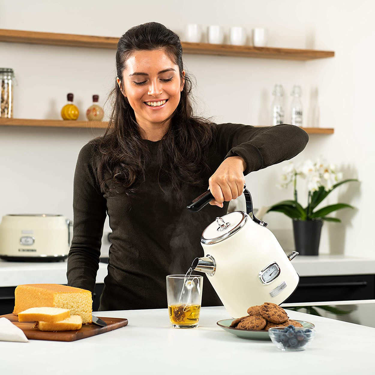 Frau gießt Tee aus einem cremefarbenen Wasserkocher in ein Glas, lächelnd.