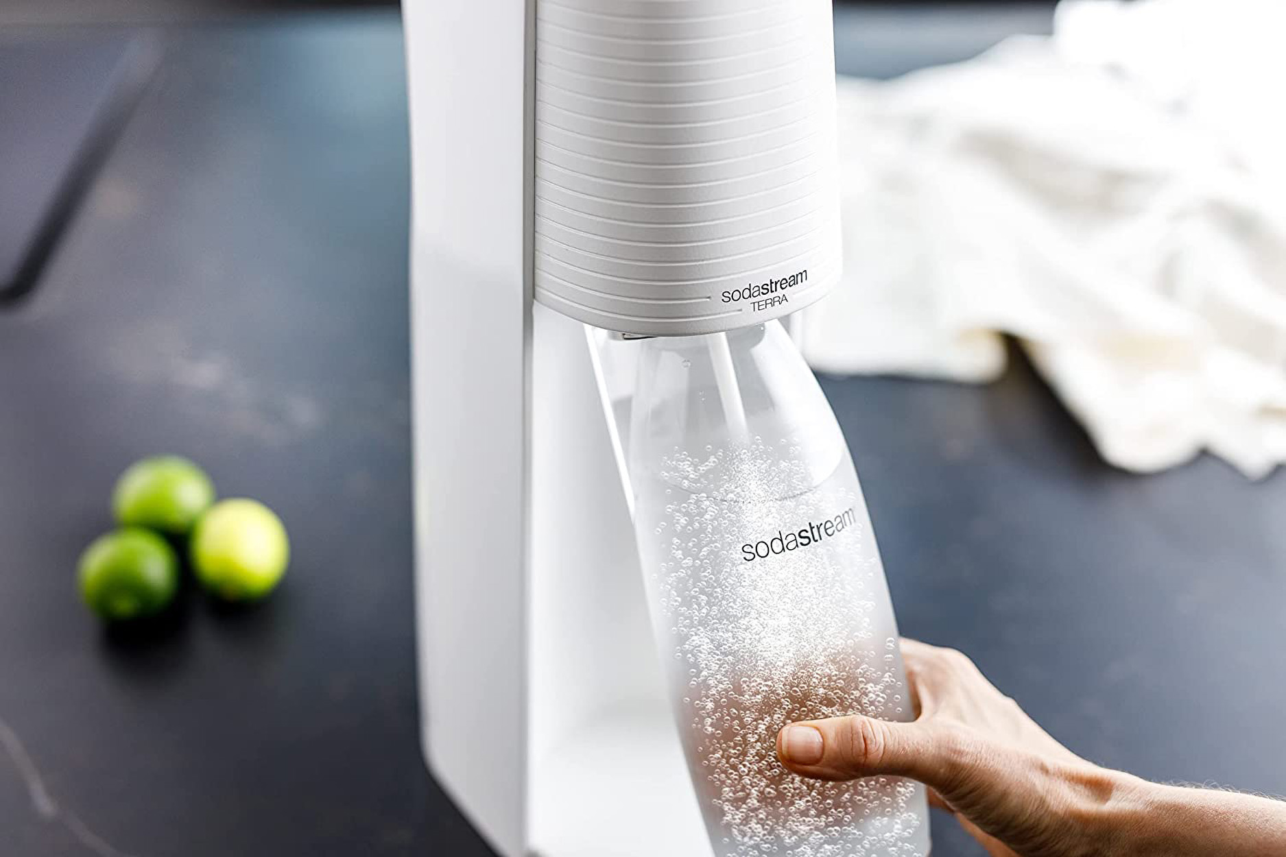 Eine Person gießt Soda aus einer weißen Sodamaschine in eine Flasche mit Limetten.
