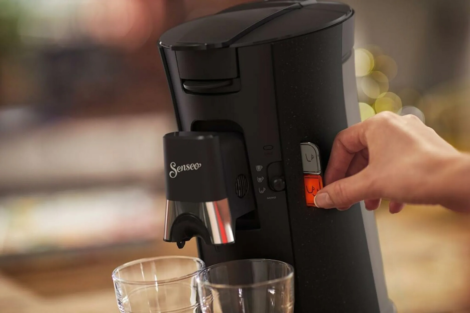 Schwarze Kaffeemaschine, Hand stellt orangefarbenes Zifferblatt neben dem Senseo-Logo ein, zwei Gläser unten.