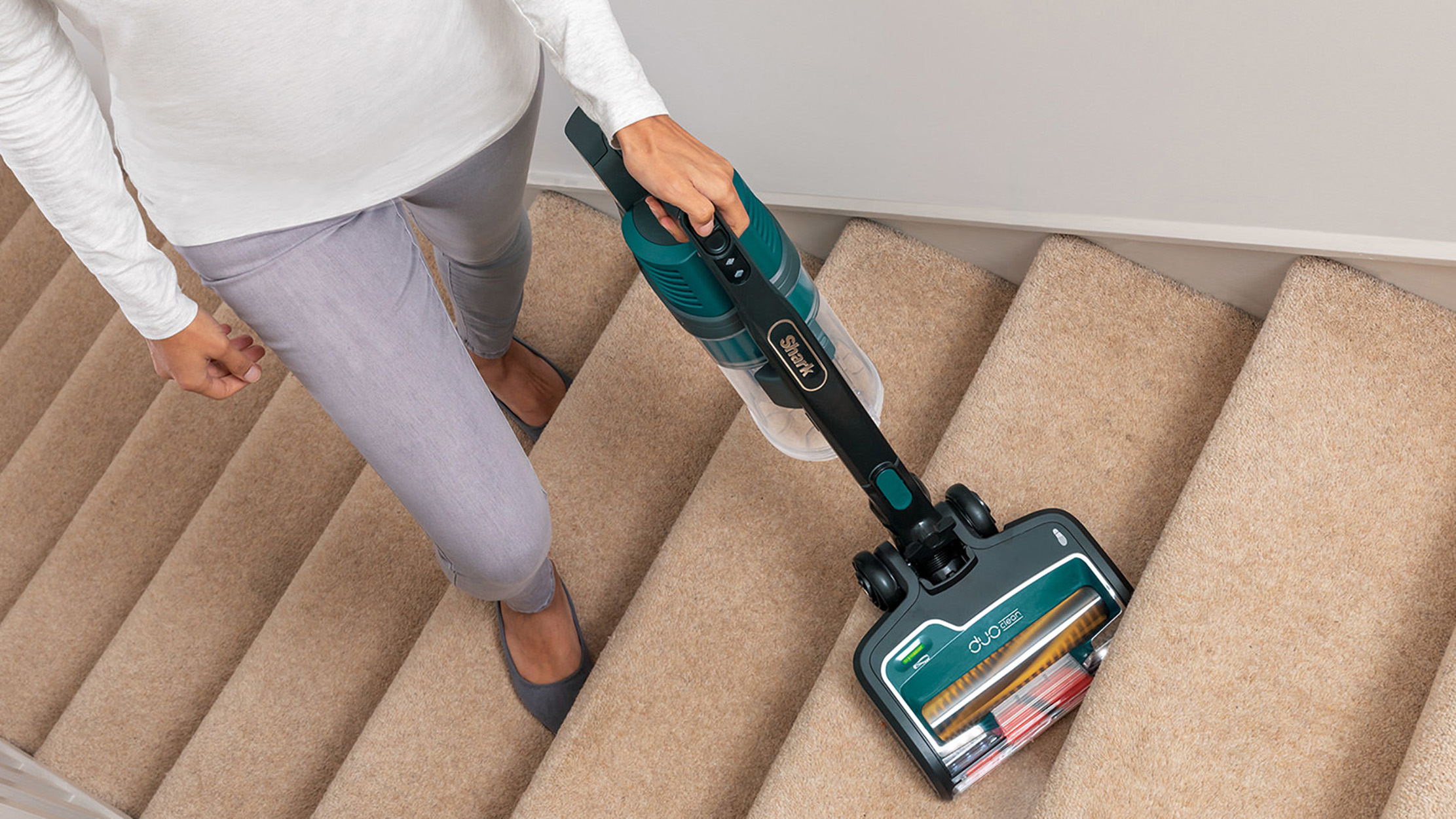 Una persona aspirando escaleras alfombradas con una aspiradora verde azulado y negra.