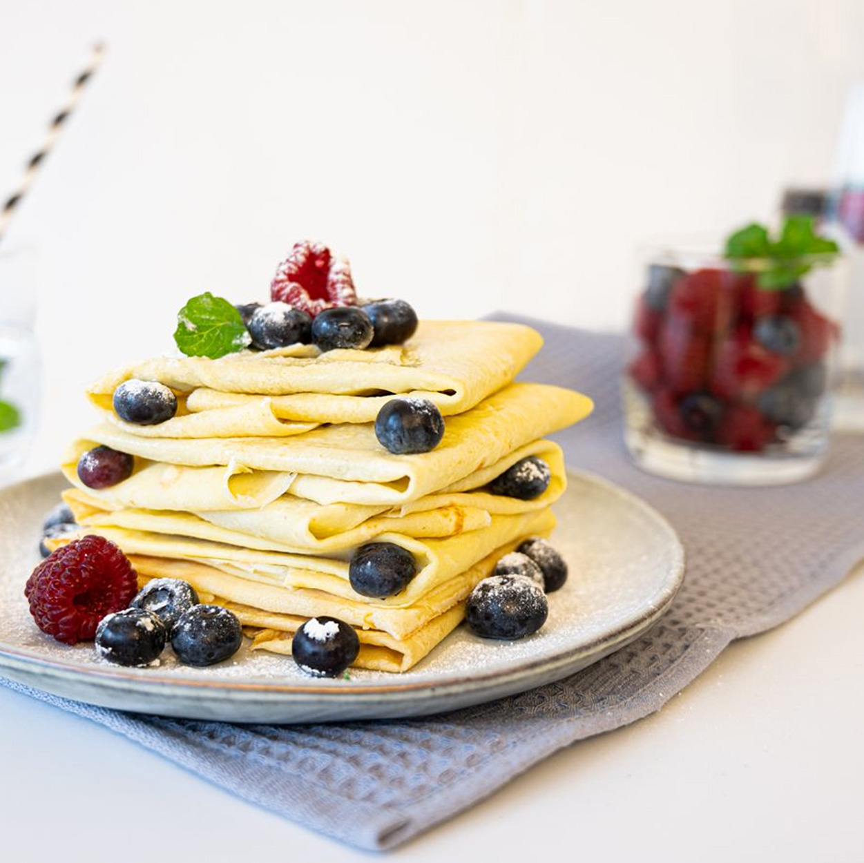 Naleśniki ułożone z jagodami i malinami na talerzu. Szklanka z jagodami.