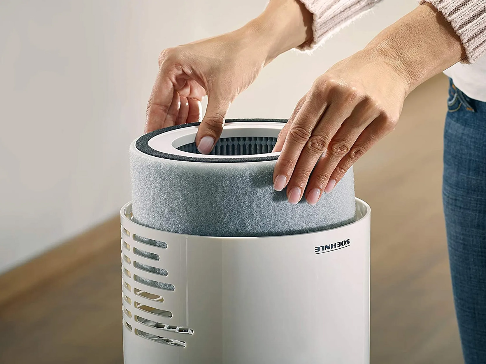 Hände legen einen Filter in einen weißen Luftreiniger ein. Grauer Filter ist sichtbar.
