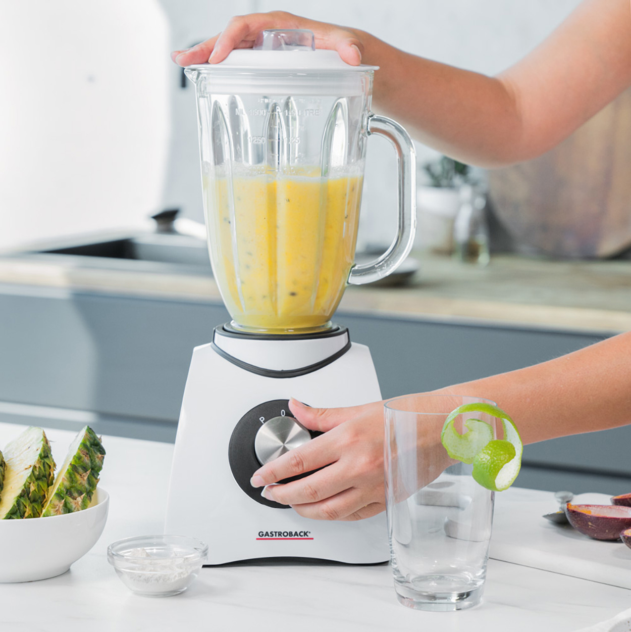 Mixer mit gelber Flüssigkeit, Ananas und Limettengarnitur, Hände und weiße Oberfläche.