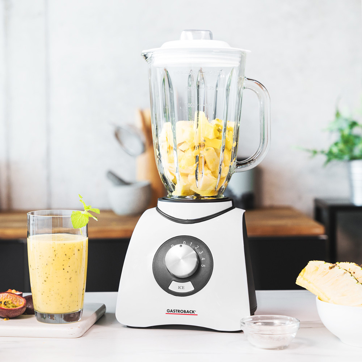 Ein Mixer mit Ananasstücken, einem Smoothie und einer Passionsfrucht auf einer Küchenzeile.