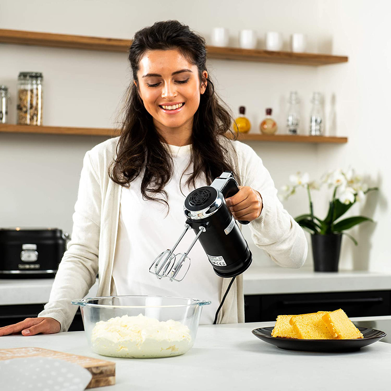 Frau, die einen schwarzen Handmixer benutzt, um Zutaten in einer Glasschüssel zu verrühren, Kuchenstücke daneben.