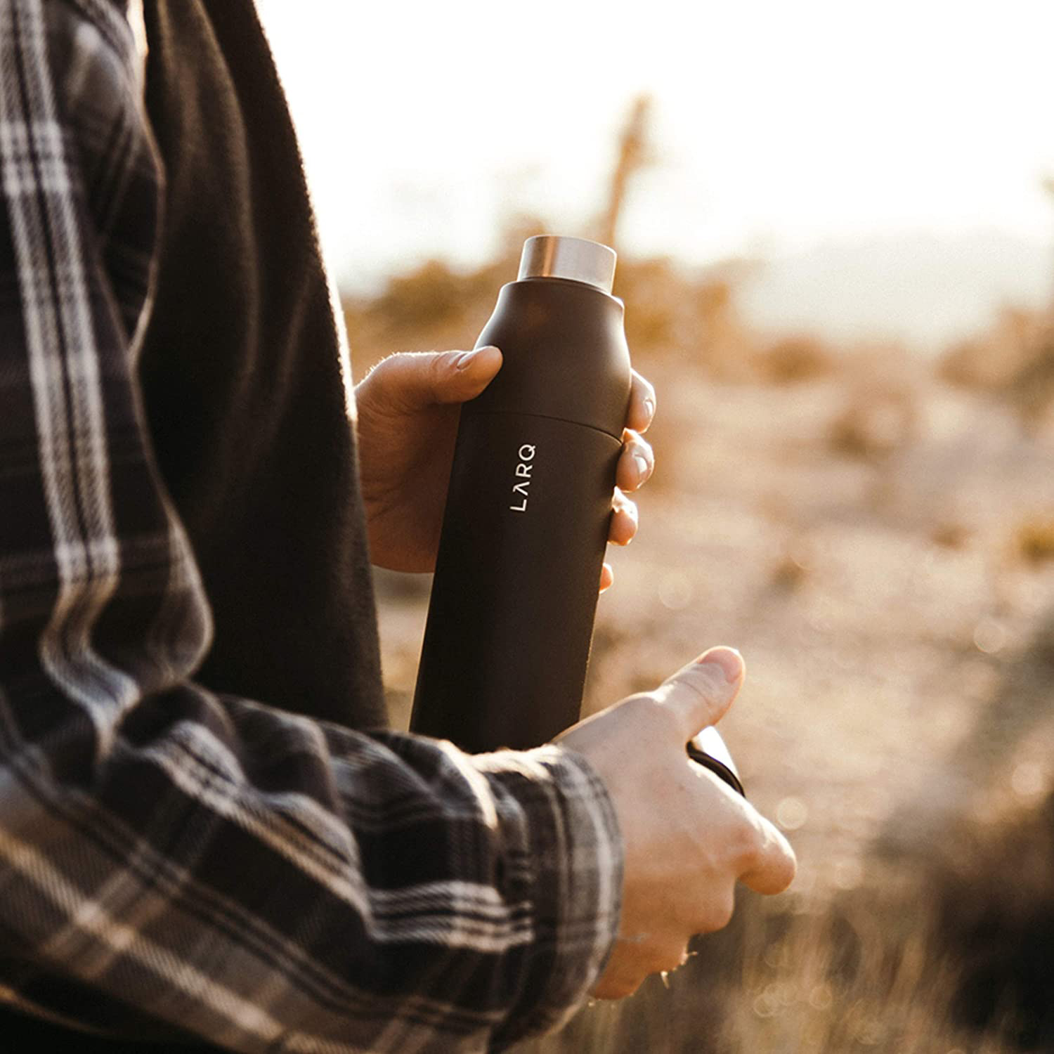 Person hält eine schwarze Wasserflasche mit silbernem Deckel in einer Wüstenumgebung.