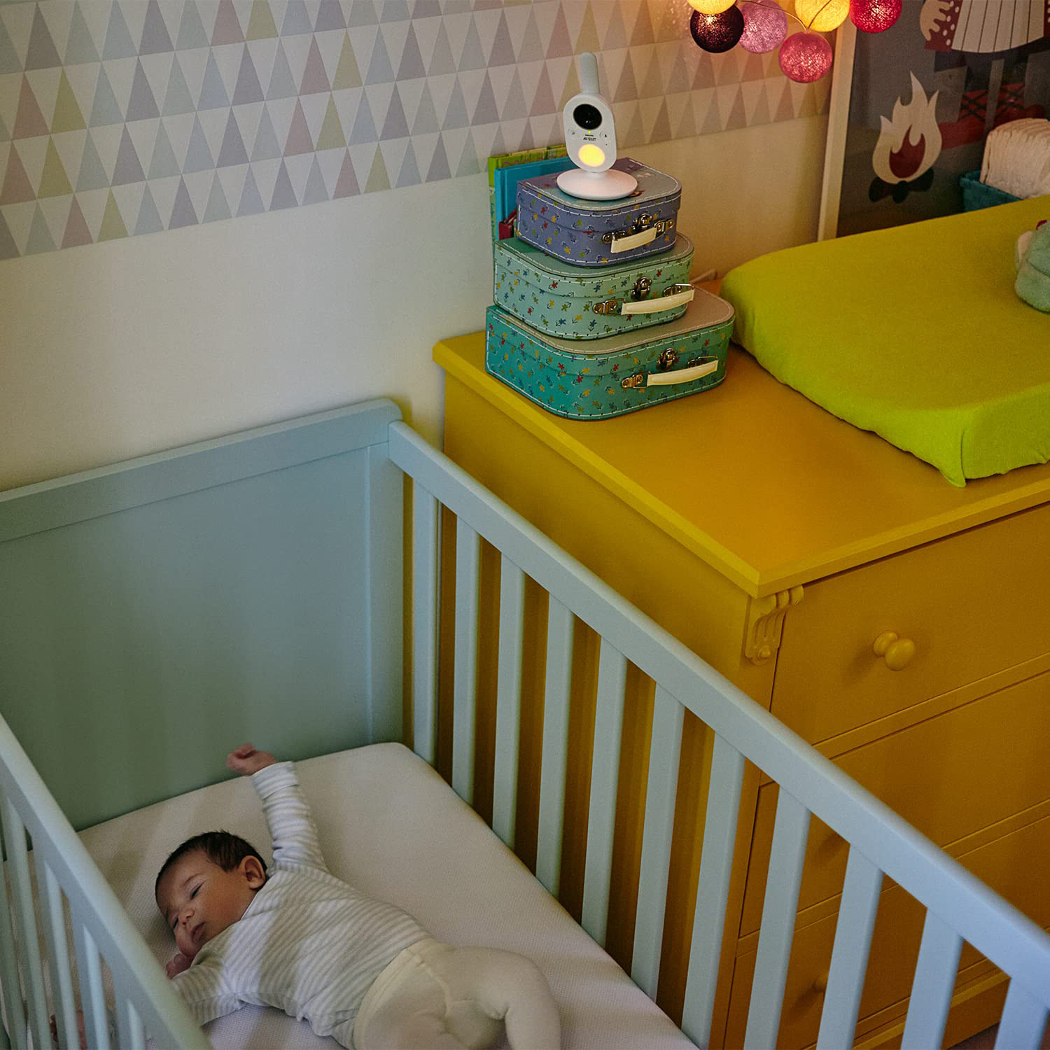 Baby schläft in blauem Kinderbett. Gelbe Kommode mit Boxen, Babyphone steht oben.