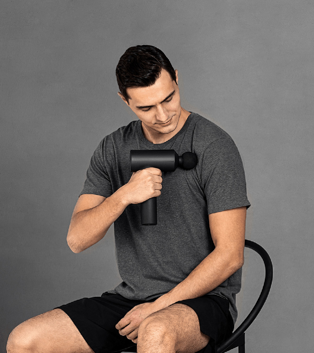 Mężczyzna używa czarnego pistoletu do masażu na ramieniu. Szare tło.