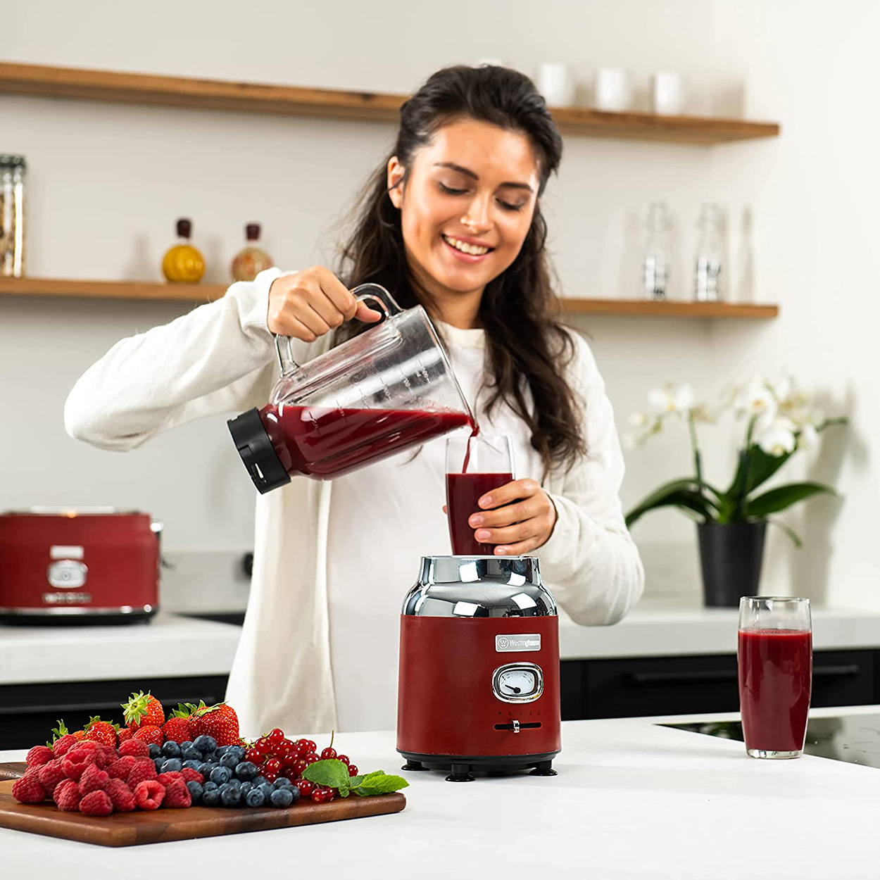Eine Frau gießt roten Saft in ein Glas. Früchte und ein Mixer sind sichtbar. Küchenumgebung.