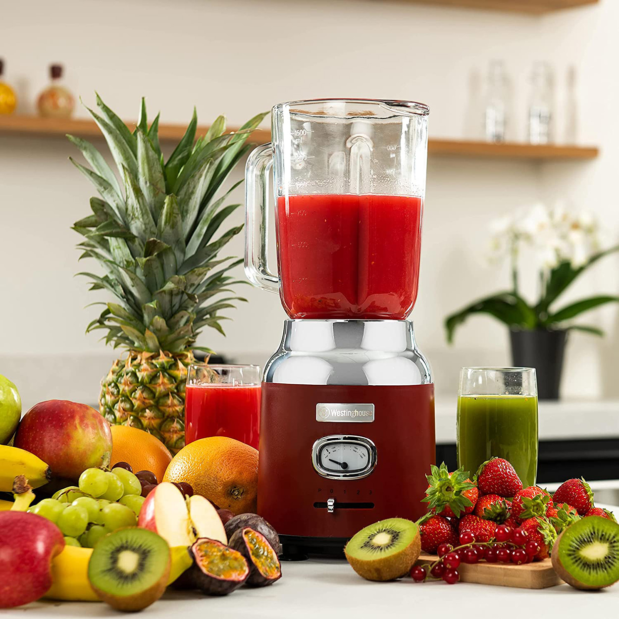 Mixer mit roter Flüssigkeit. Früchte, darunter Ananas und Kiwi, sind in der Nähe.