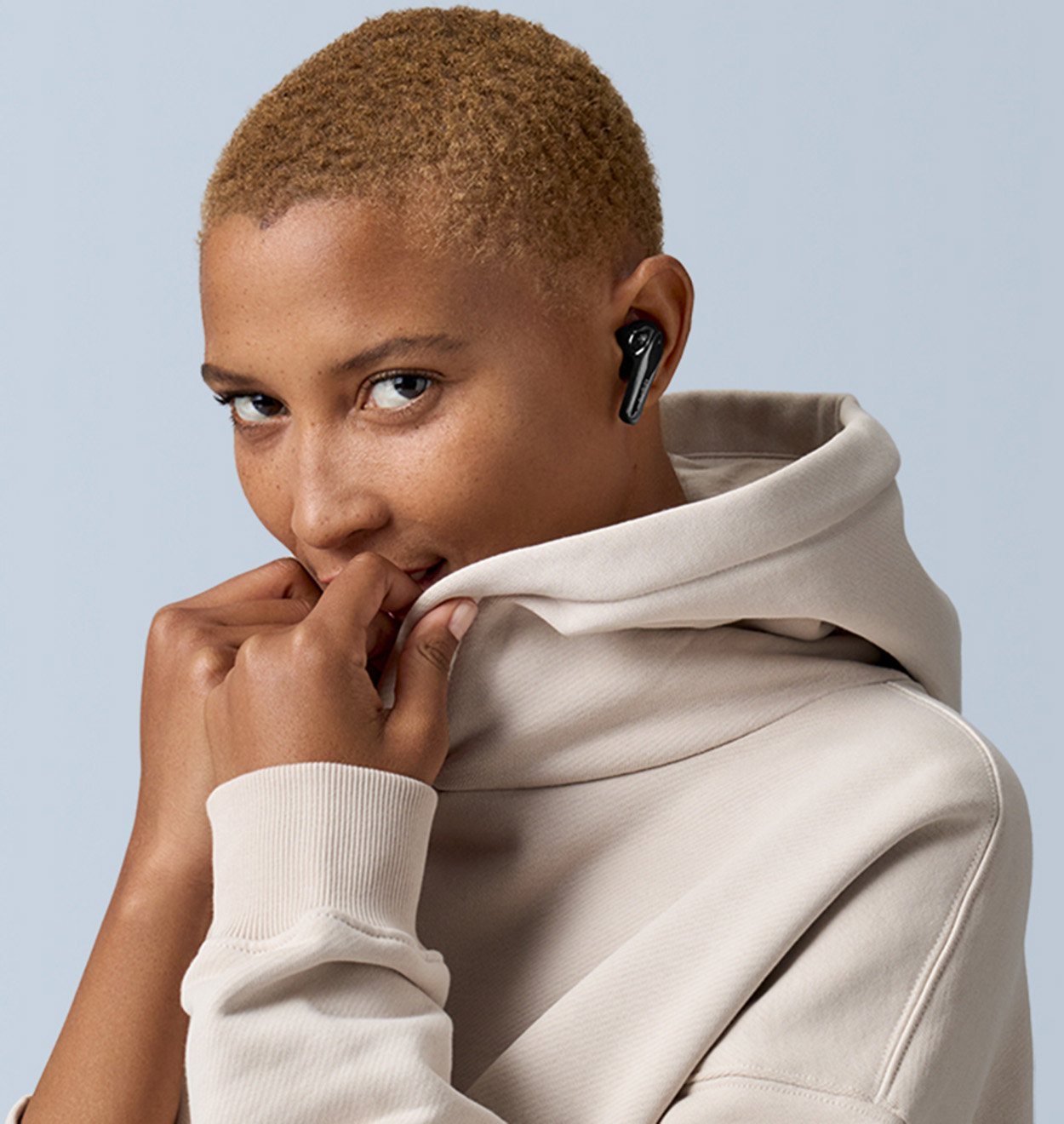 Kobieta w beżowej bluzie z kapturem, trzymająca kołnierz rękami, nosząca czarne słuchawki.