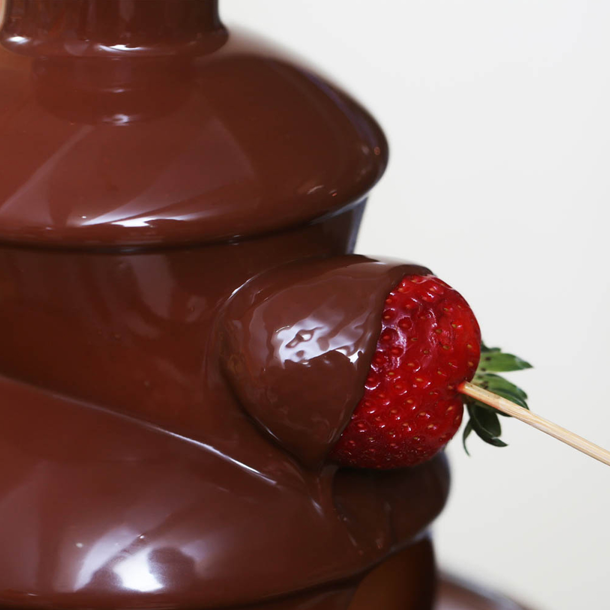 Fresa sumergida en chocolate, sujeta por un palillo, con fuente de chocolate detrás.