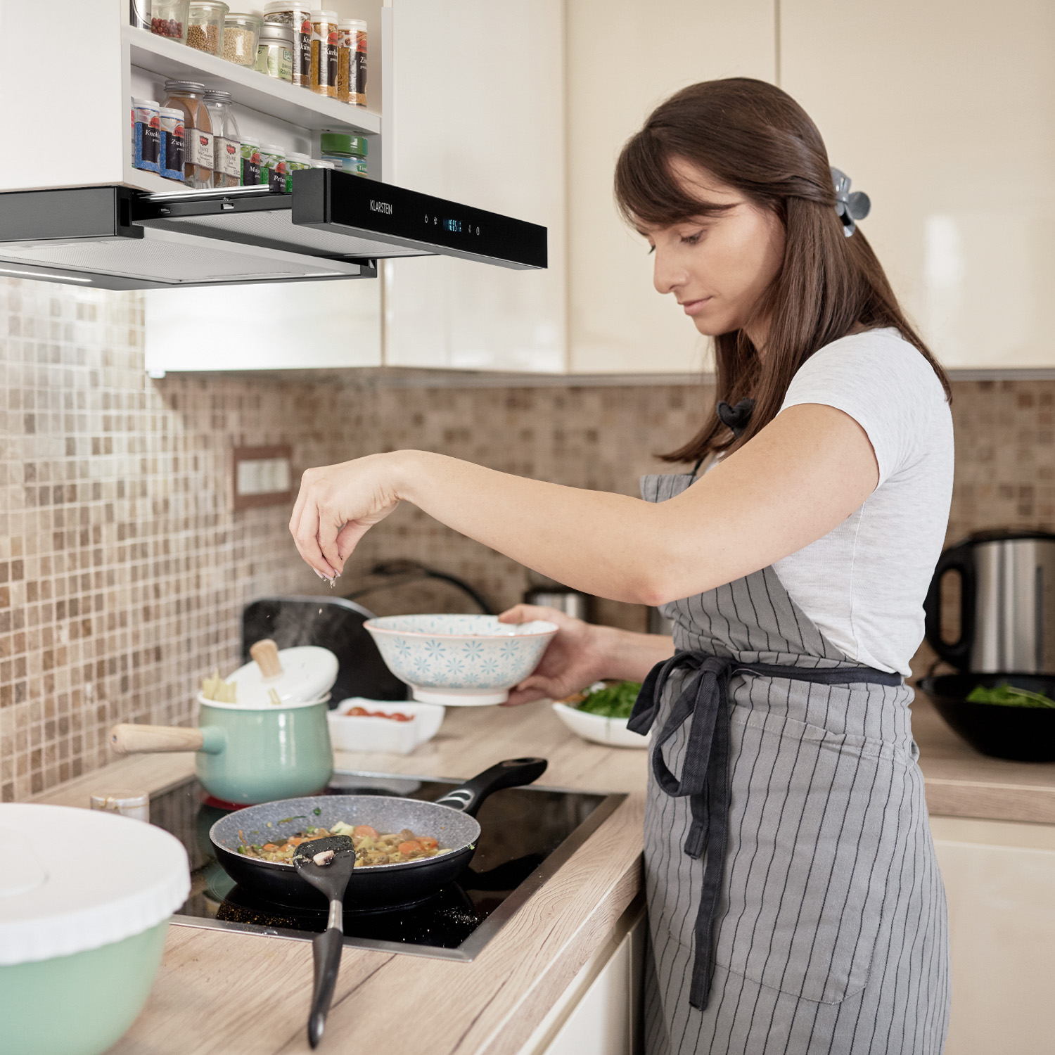 Frau in der Küche beim Kochen, streut Gewürze in eine Pfanne. Dunstabzugshaube darüber.