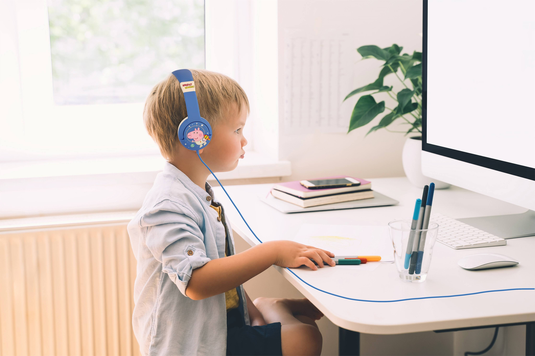 Chłopiec z niebieskimi słuchawkami Peppa Pig patrzy na ekran komputera, siedząc przy biurku.