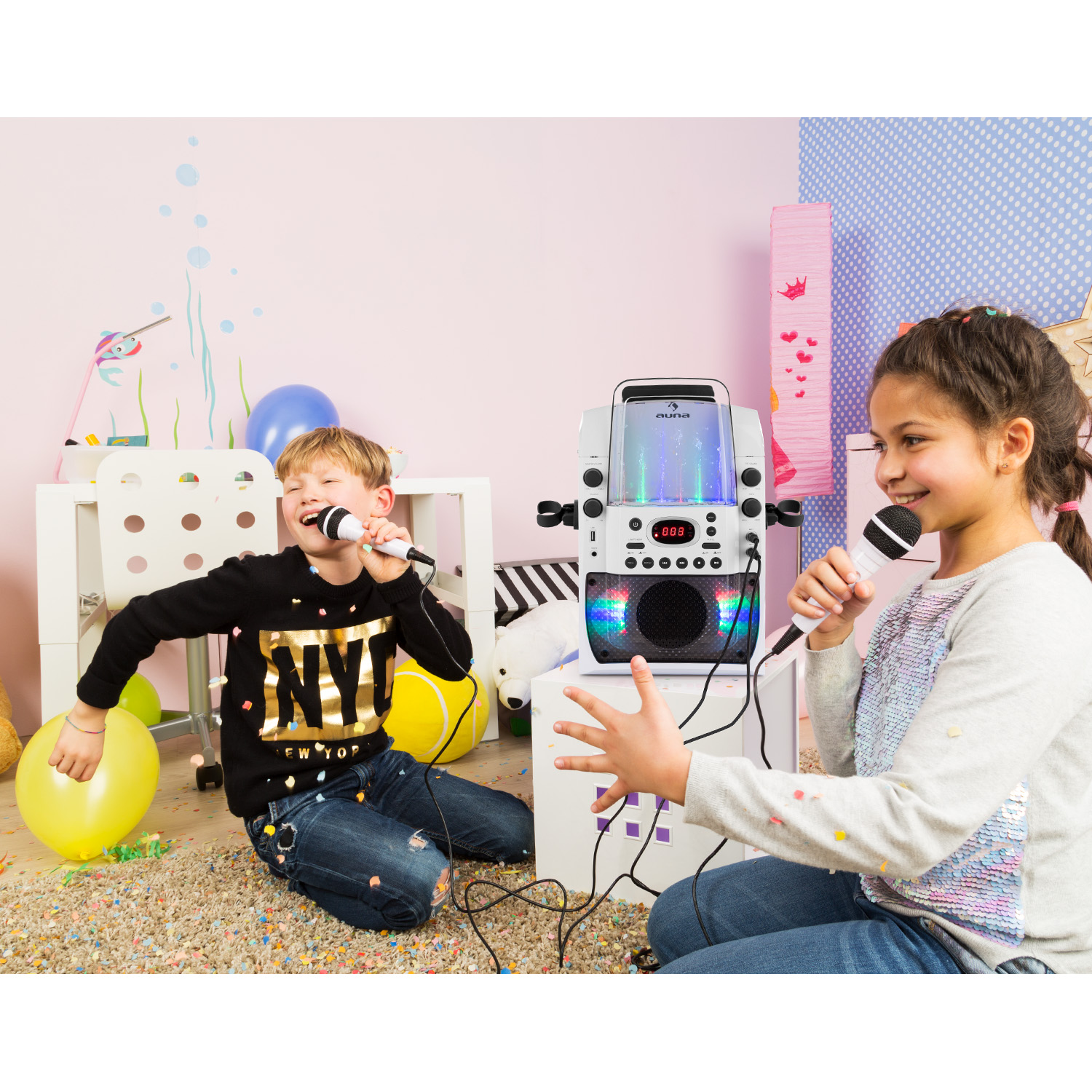 Zwei Kinder singen in Mikrofone neben einer Karaoke-Maschine in einem farbenfrohen Raum.