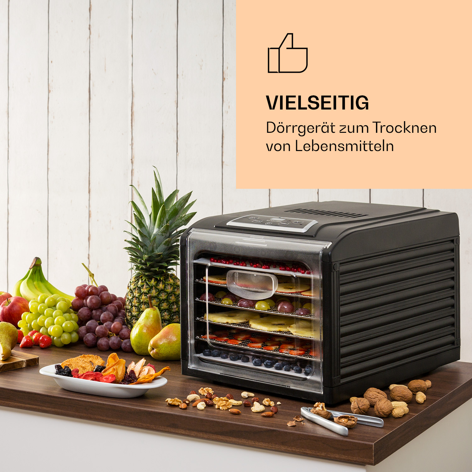 Ein schwarzer Dörrautomat mit Essen drin, Früchte und Nüsse auf dem Tisch.