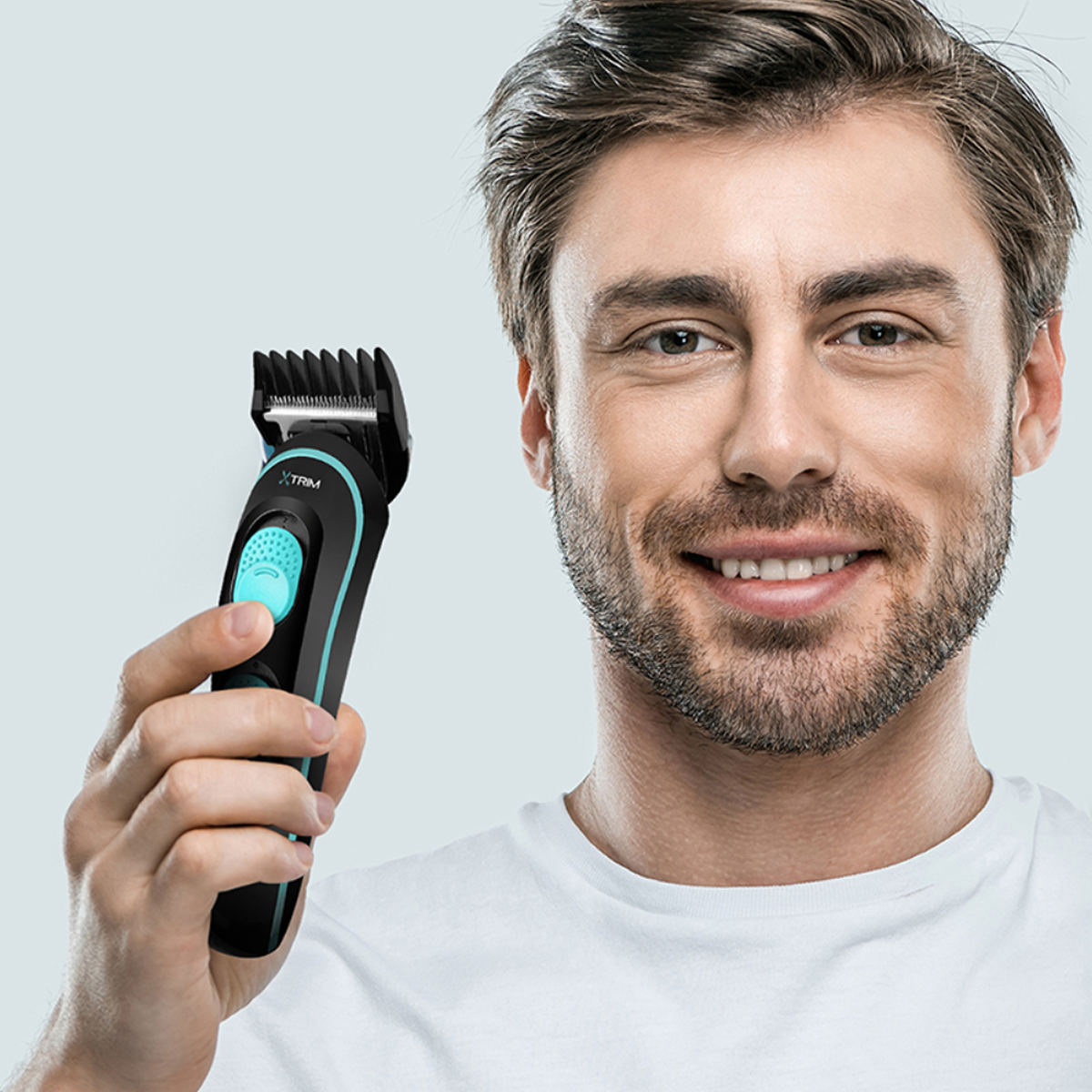 Hombre sosteniendo un recortador eléctrico negro y verde azulado. Sonriendo, camiseta blanca. Fondo azul claro.