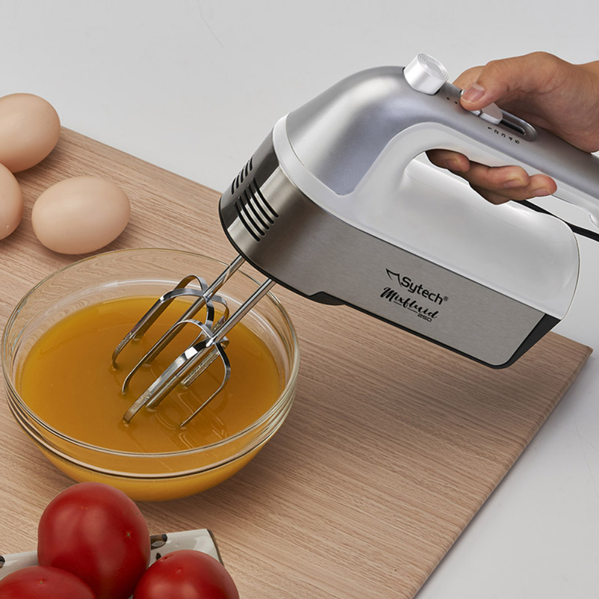 Batidora de mano mezclando líquido en un bol. Huevos, tomates y una tabla de madera en la escena.