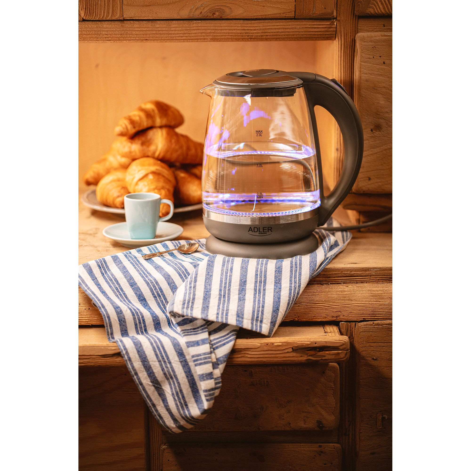 Bouilloire électrique en verre avec croissants, tasse et serviette sur bois.