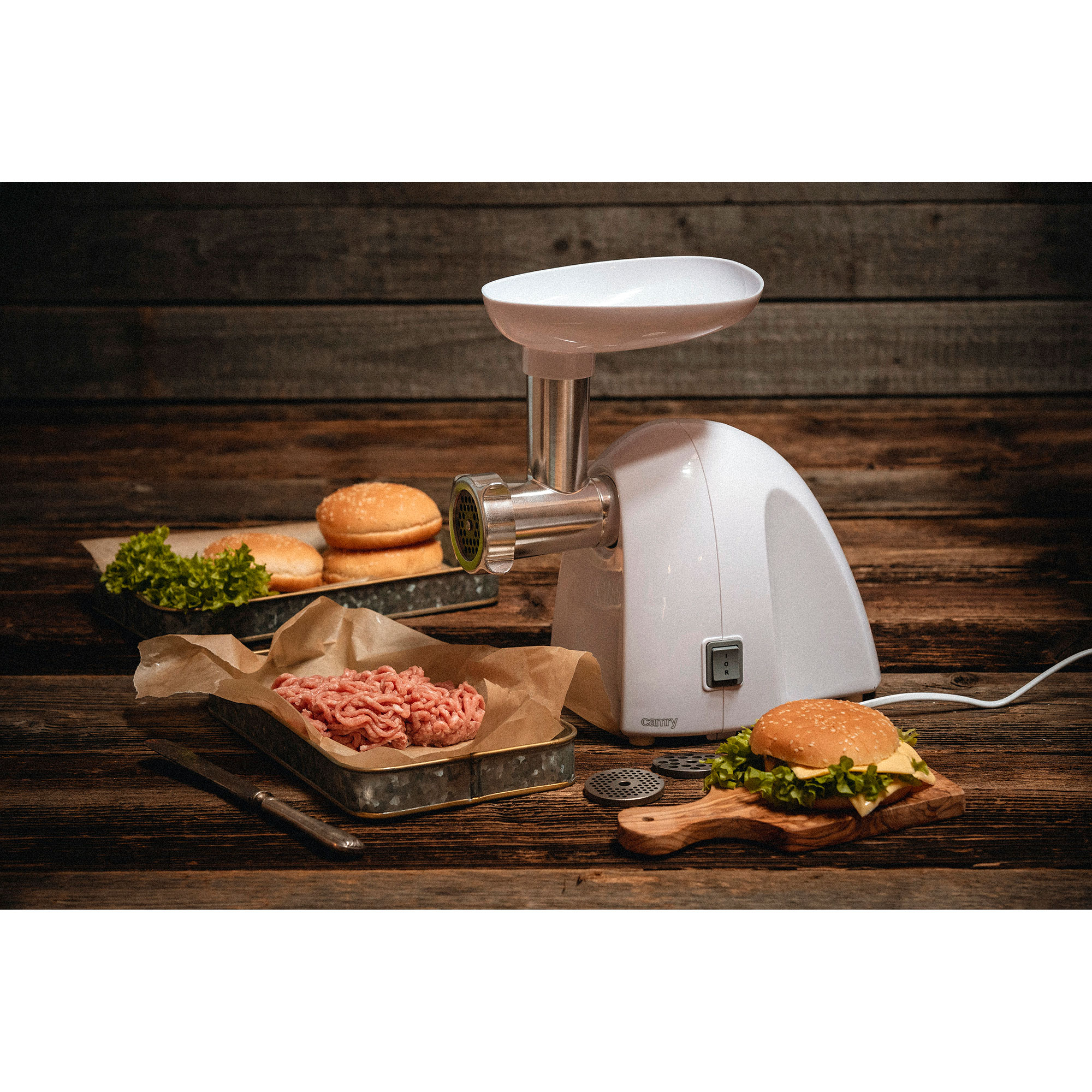 Biała maszynka do mielenia mięsa na drewnianym stole, z przygotowaniem jedzenia i burgerami. Ciemne drewniane tło.
