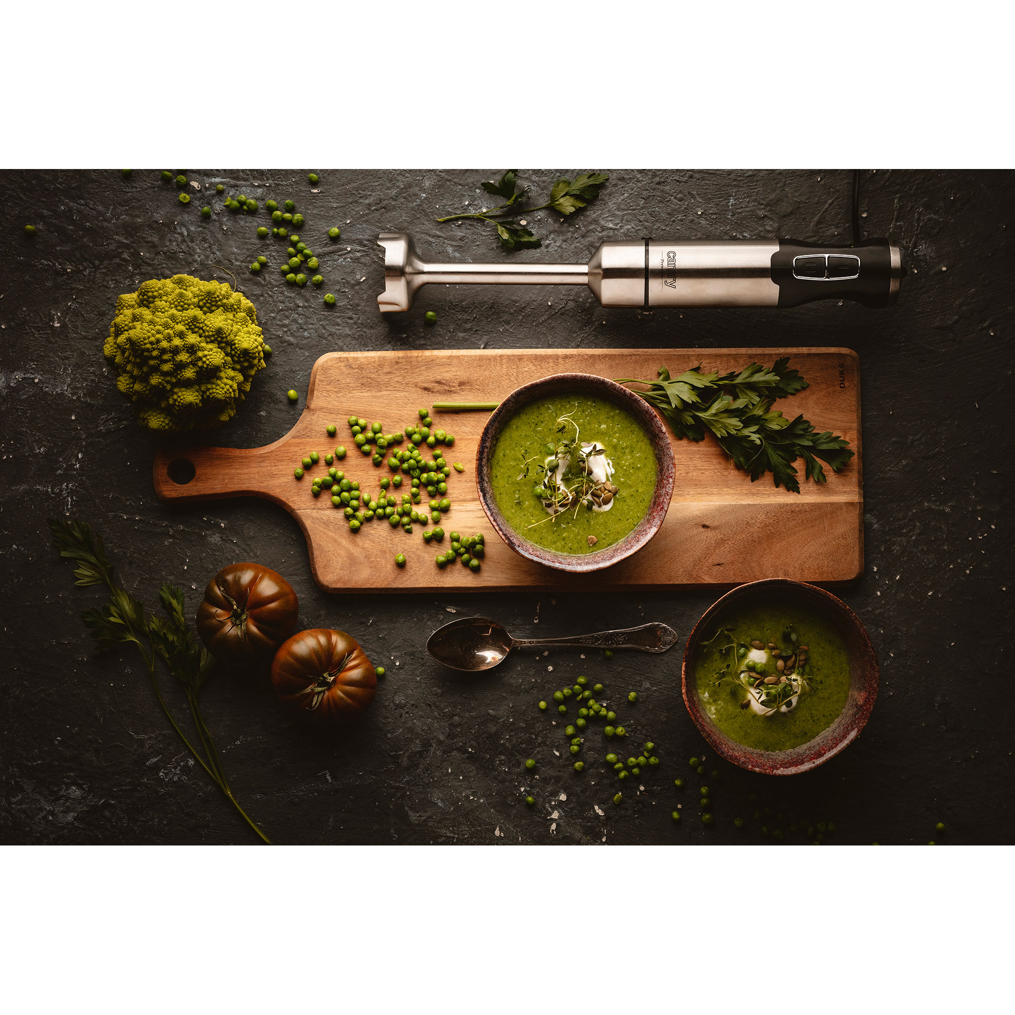 Zupa w miskach na drewnianej desce. Widoczne są również groszek, zioła i blender ręczny.