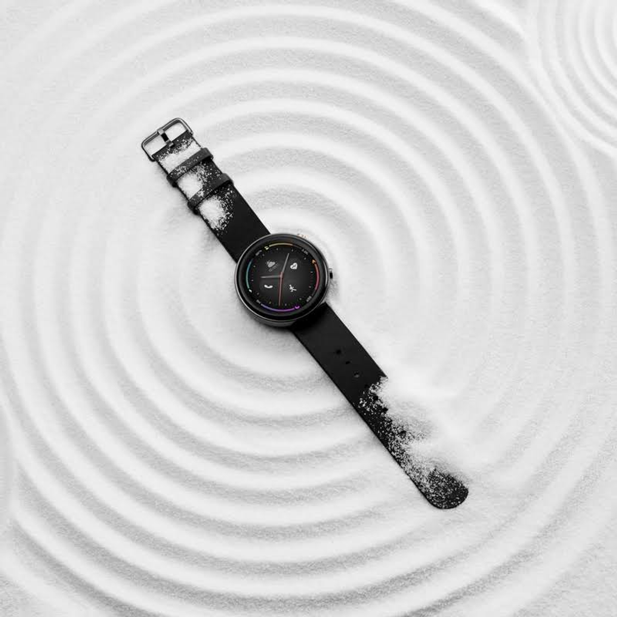 Smartwatch negra sobre arena blanca con círculos concéntricos, correa de reloj negra.