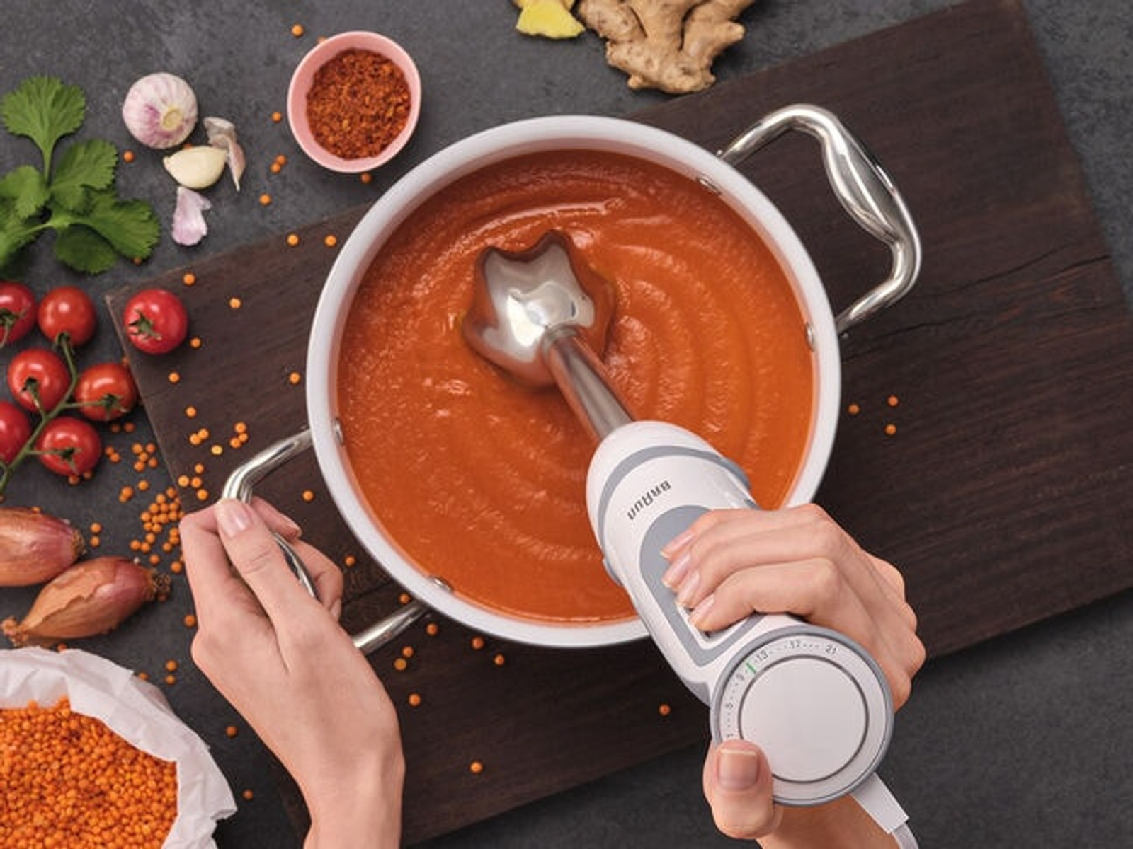 Turuncu çorba dolu bir tencerede daldırma blender kullanan eller. Tencerenin etrafına serpilmiş malzemeler.