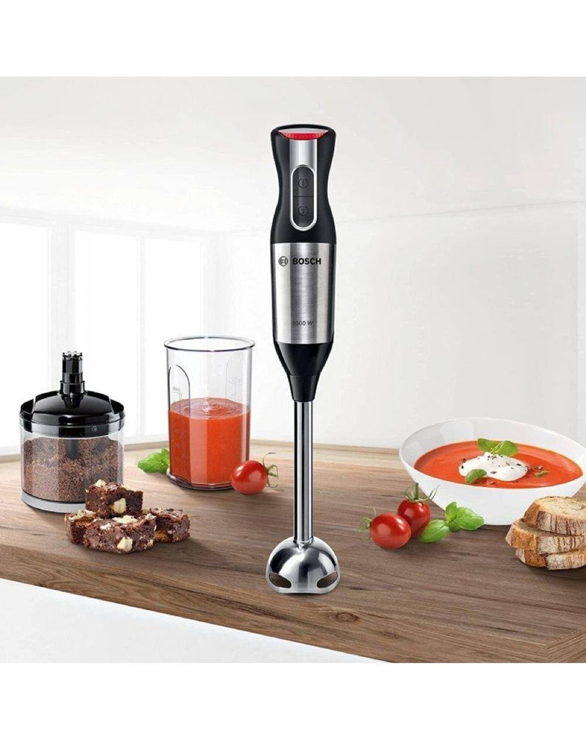 Frullatore a immersione con accessori. Include frullatore, bicchiere, pomodori, pane e brownies. Superficie in legno.