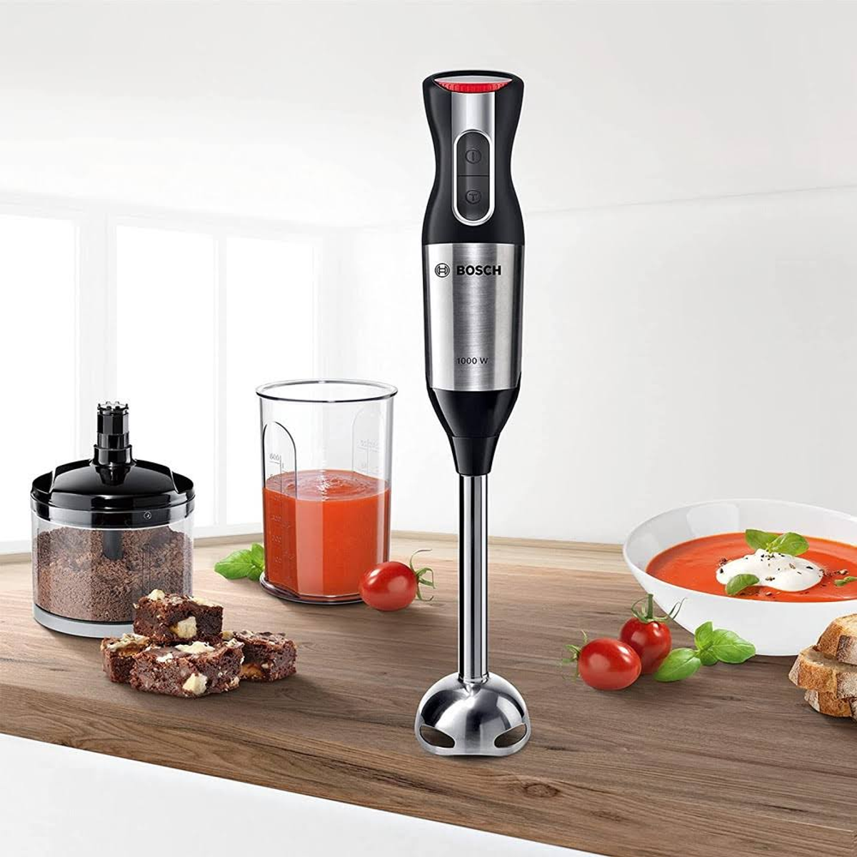 Frullatore a immersione con accessori. Un bicchiere di zuppa di pomodoro, pomodori e brownies sono sul tavolo.
