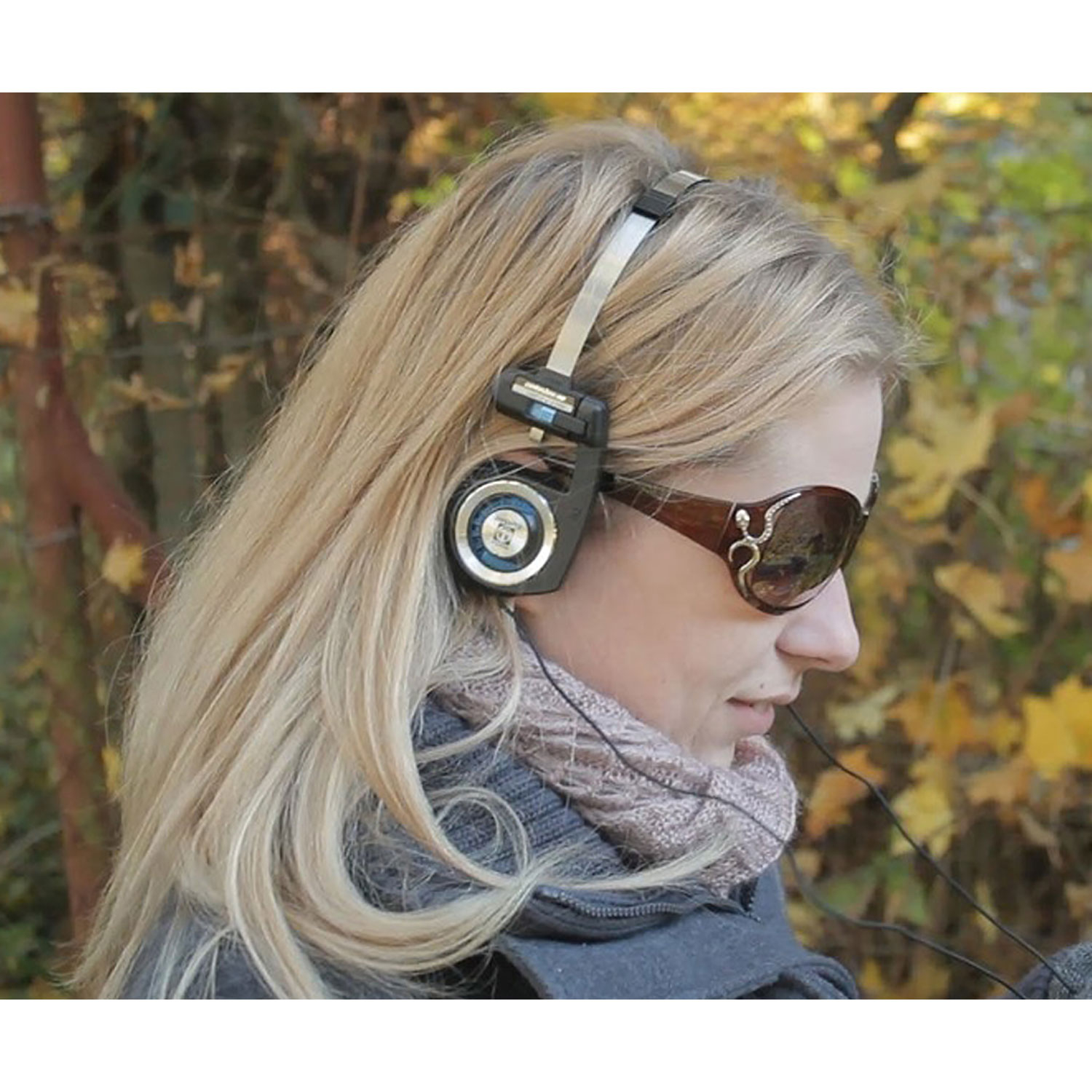 Una mujer rubia con auriculares y gafas de sol. Está al aire libre con hojas de otoño en el fondo.