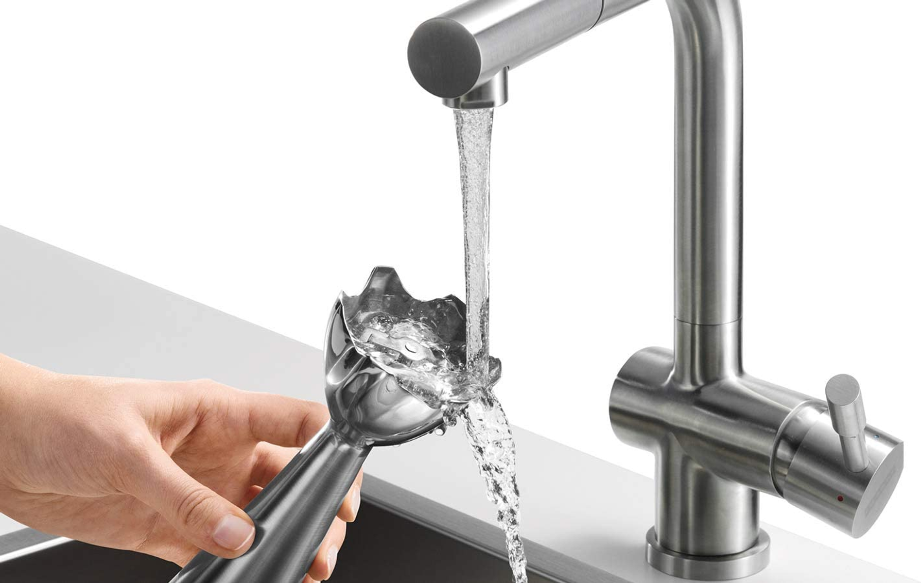 Mano che tiene una testa di frullatore sotto un rubinetto. L'acqua scorre. Sfondo bianco.