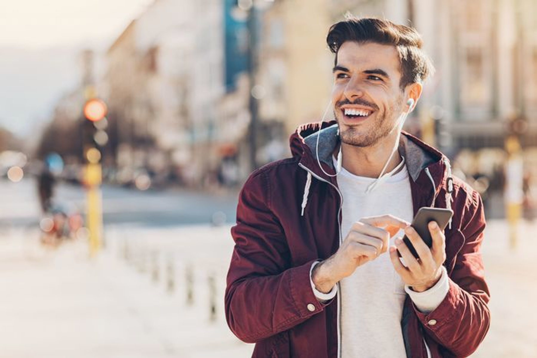 Uomo con giacca bordeaux, usa il telefono con auricolari per strada. Sorridente e felice.