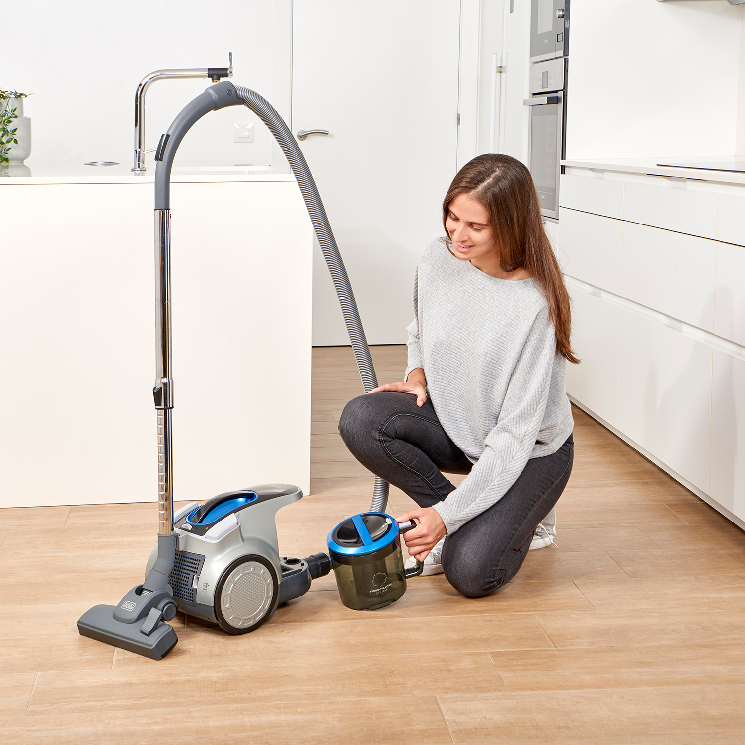 Kobieta klęczy obok odkurzacza, trzymając pojemnik na kurz w kuchni.