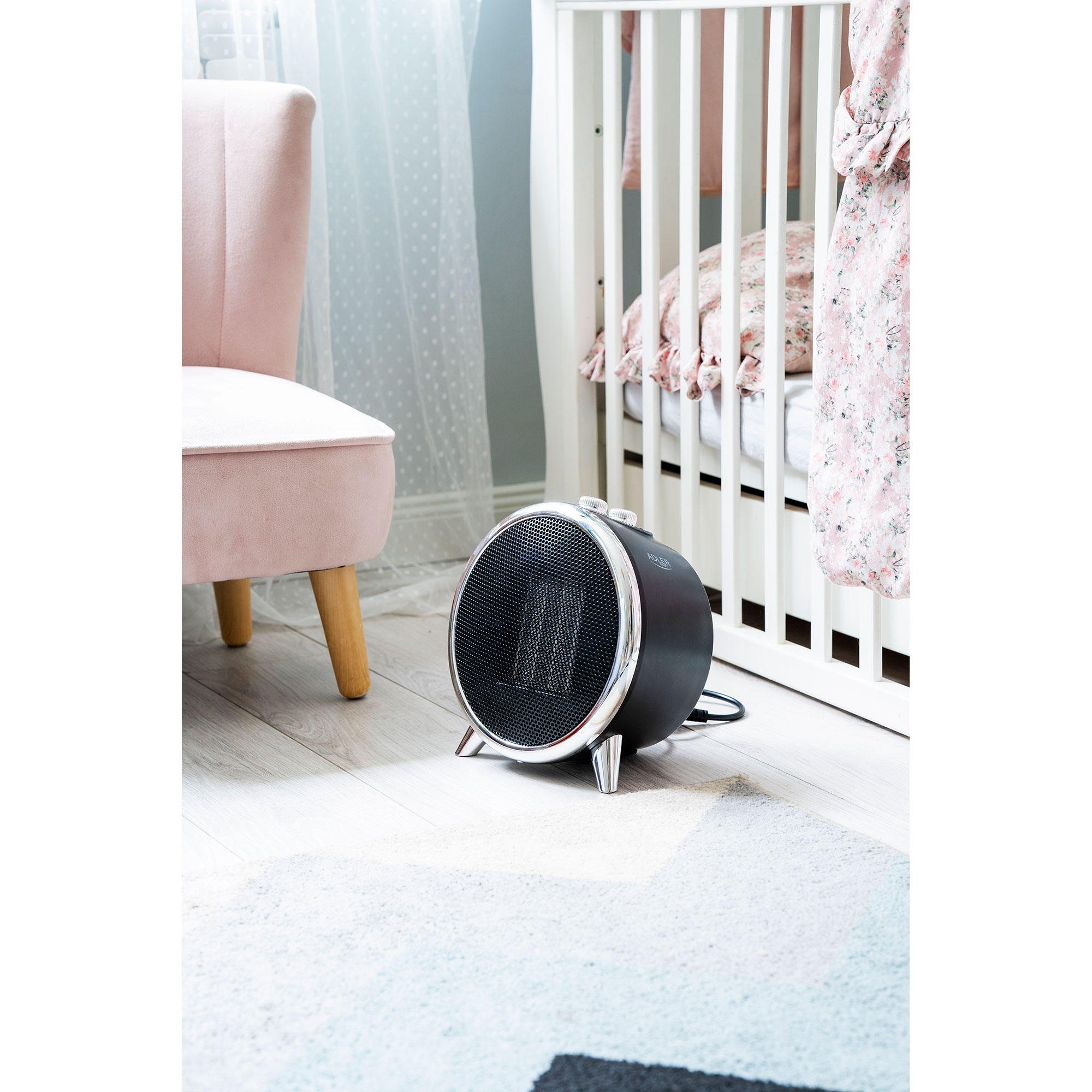Czarny grzejnik elektryczny w pokoju. Różowe krzesło po lewej, białe łóżeczko w tle.