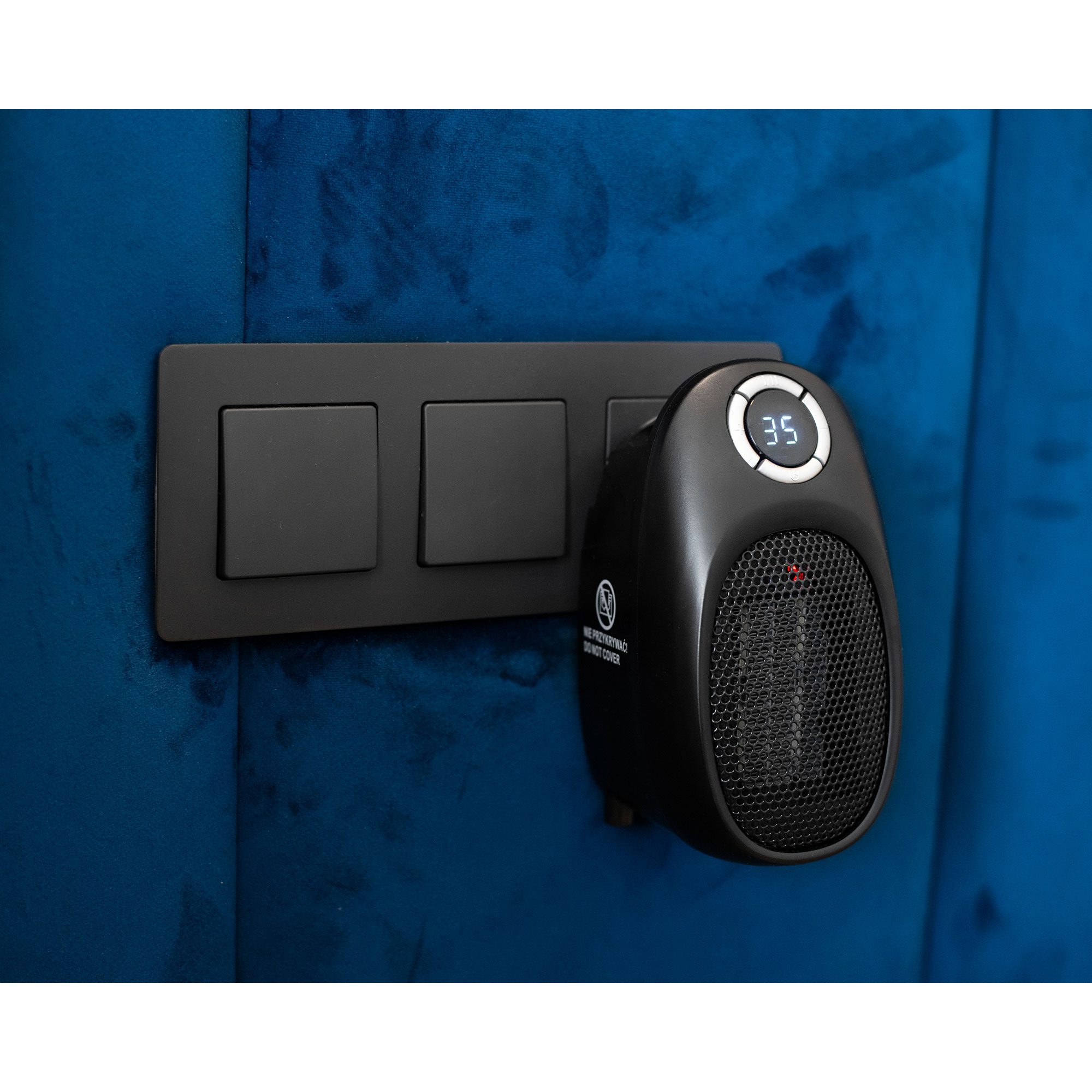 Ein schwarzer Heizlüfter mit digitaler Anzeige und eine schwarze Schalttafel an einer blauen Wand.