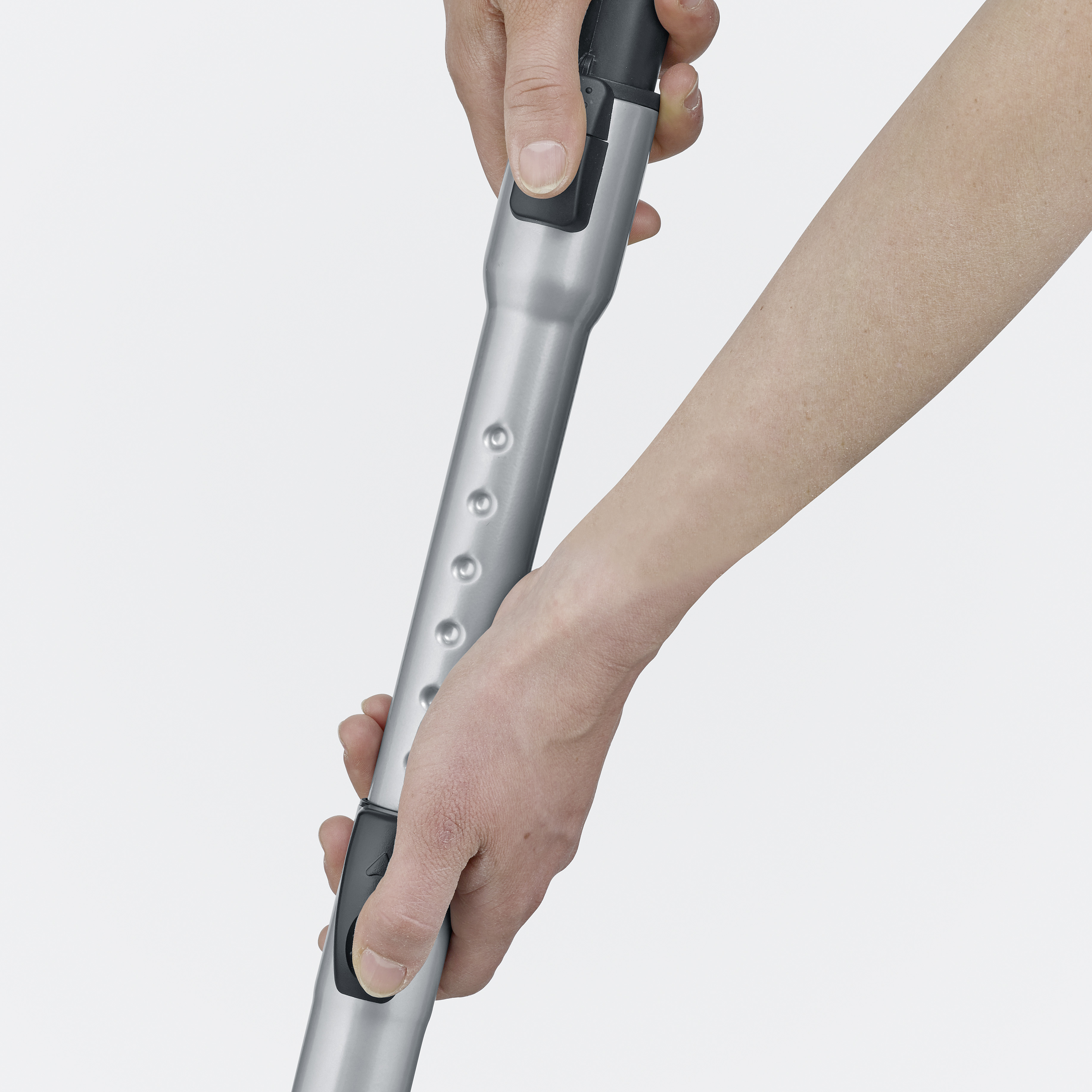 Dłonie chwytające srebrną rurę przedłużającą do odkurzacza. Tło jest białe.