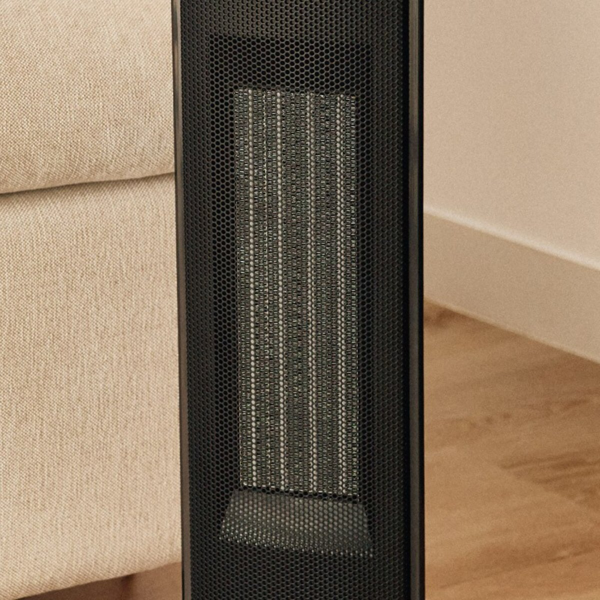Primer plano de un calefactor eléctrico negro con rejilla de panal y sofá beige detrás.