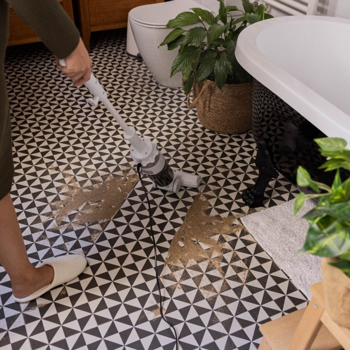 Mujer aspirando un suelo de baldosas estampadas. Ambiente de baño. Bañera y plantas visibles.