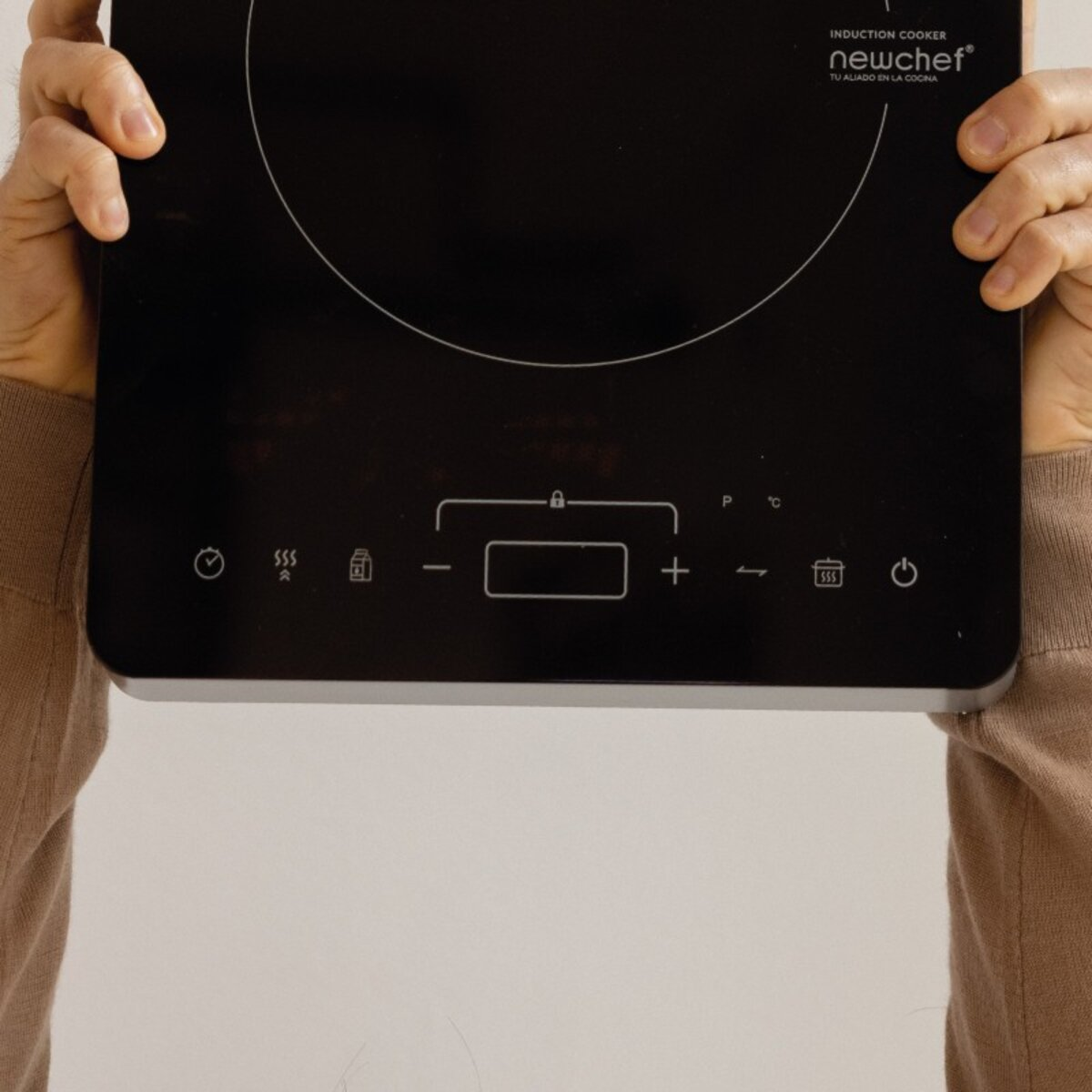 Cocina de inducción negra. Manos la sostienen. Botones y símbolos en la superficie. Fondo blanco.