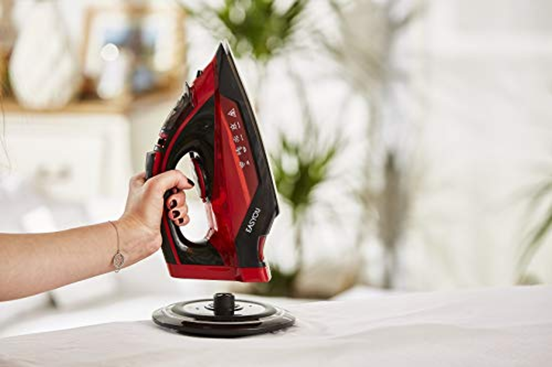 Hand hält rotes und schwarzes Bügeleisen über weißem Tuch, schwarze Basis daneben, Hintergrund.